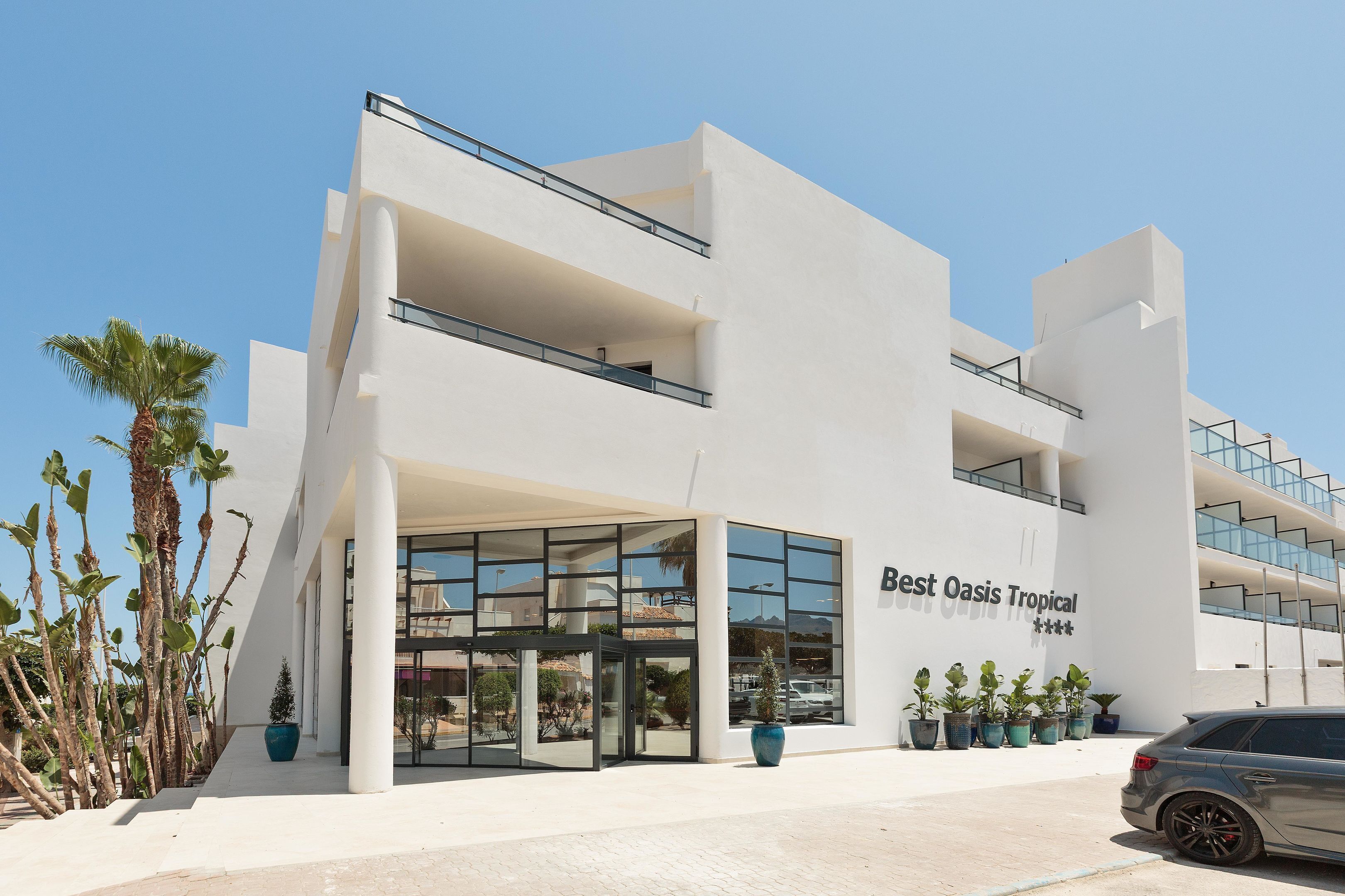 A modern building with a sign reading "Best Oasis Tropical." It features a glass entrance, multiple balconies, and potted plants around the entrance.
