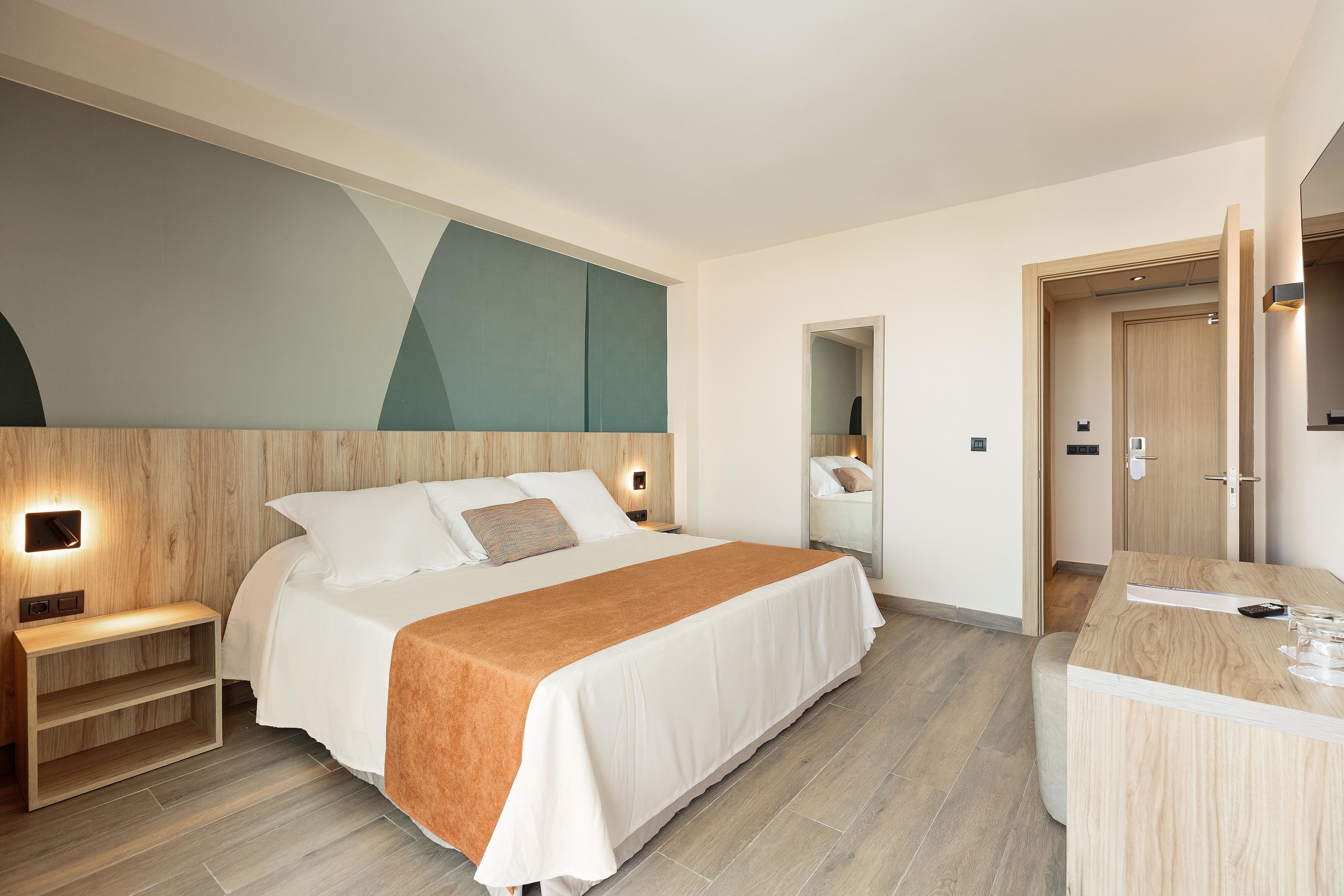 A modern hotel room with a large bed, bedside tables, a wooden desk, and a mirror.