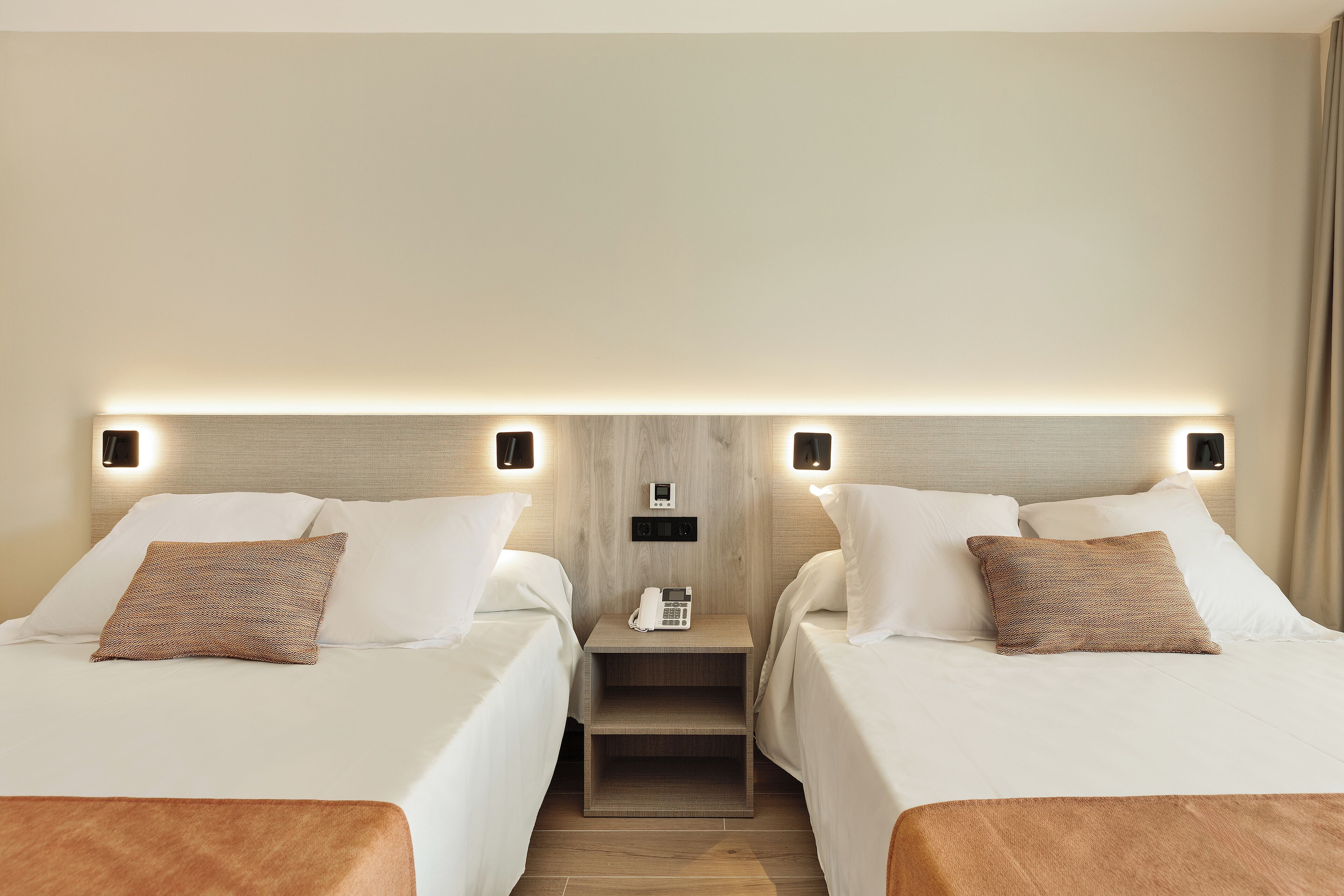 A hotel room with two beds, each with white linens and brown pillows. There is a nightstand between the beds with a phone and a small table. The room has a wooden headboard and a light fixture above the nightstand.