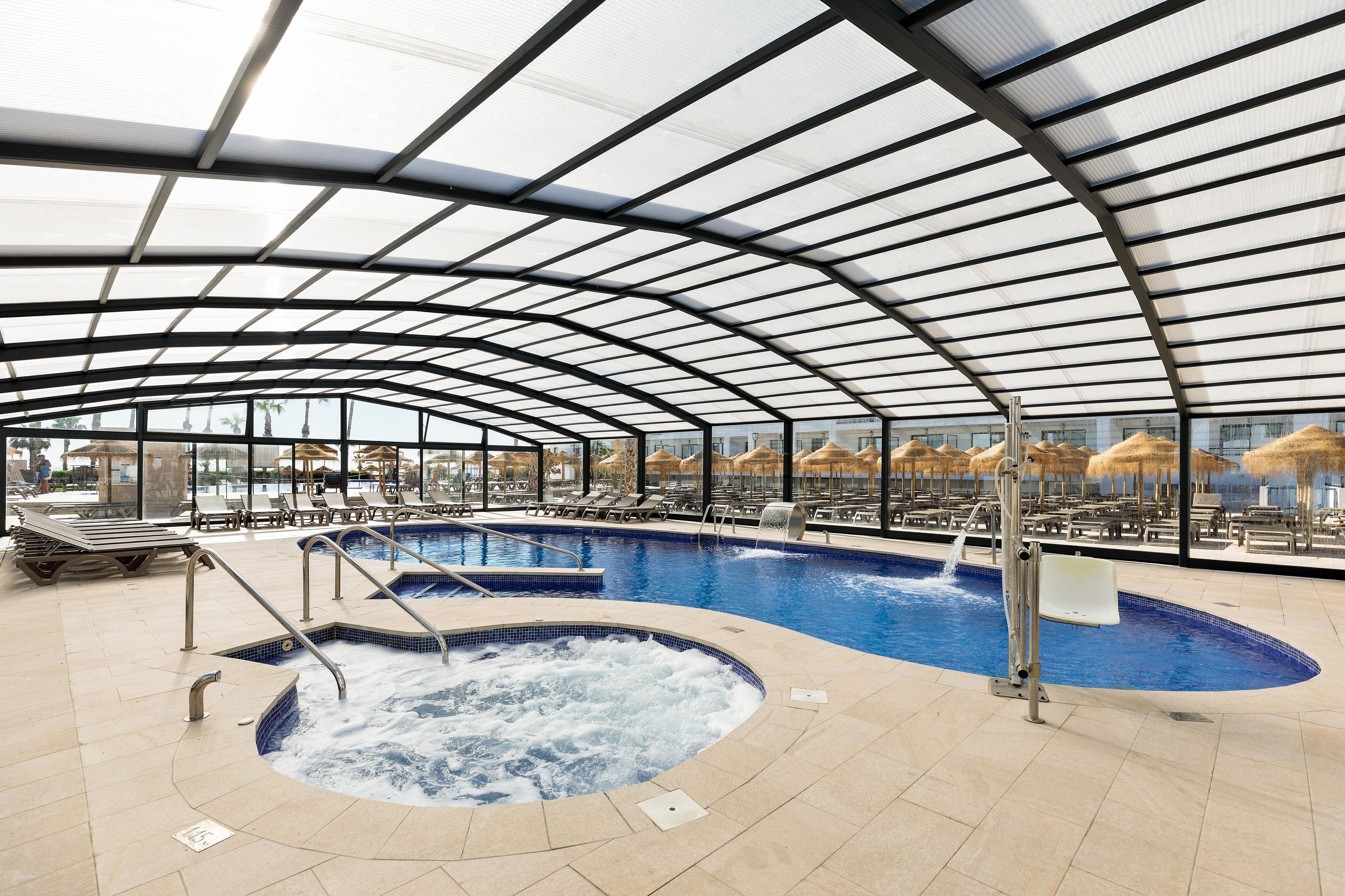 An indoor swimming pool with a glass ceiling, lounge chairs, and a hot tub.