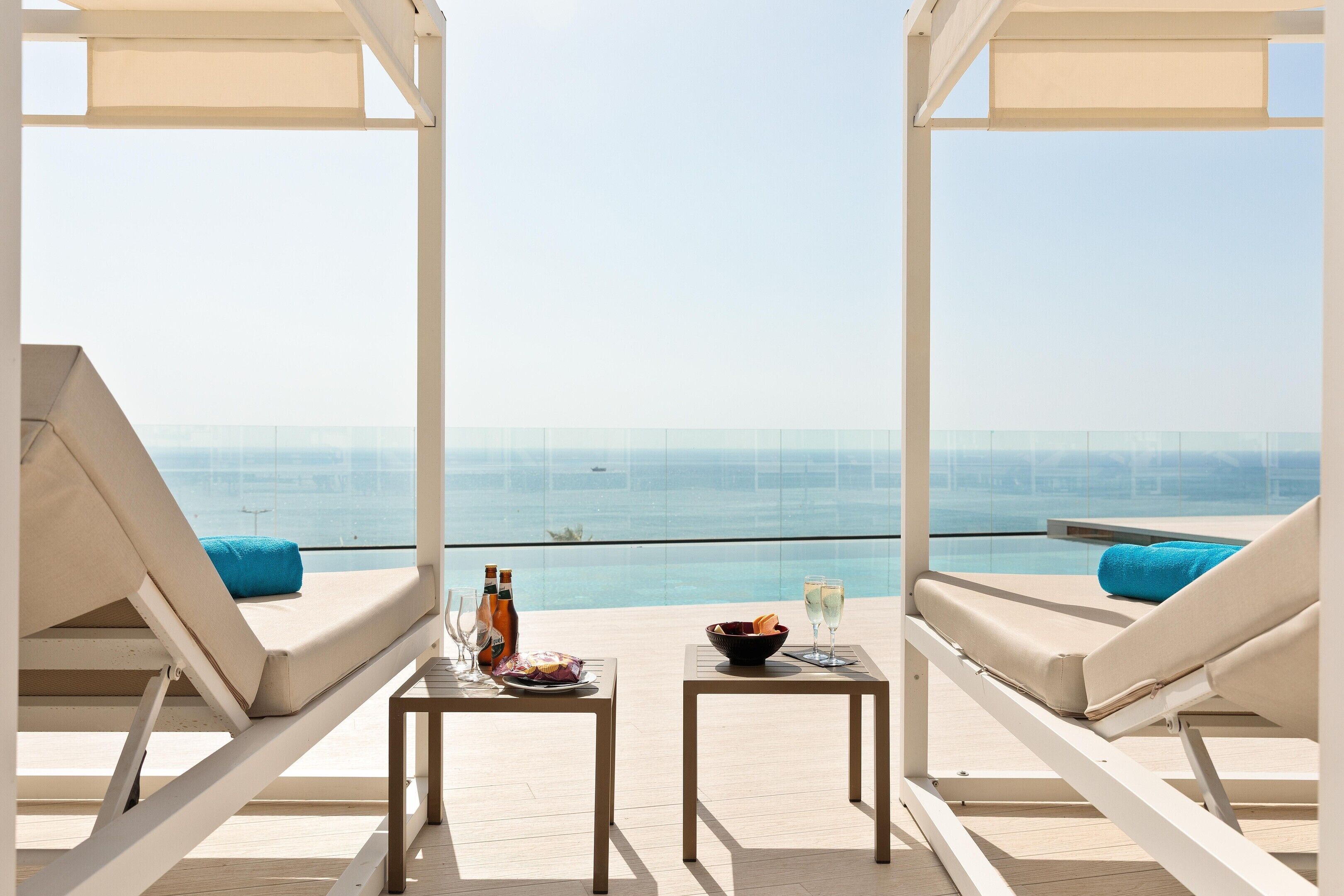 Un área de estar al aire libre iluminada por el sol, con dos sillones, una mesa con bebidas y aperitivos, y vista al mar.