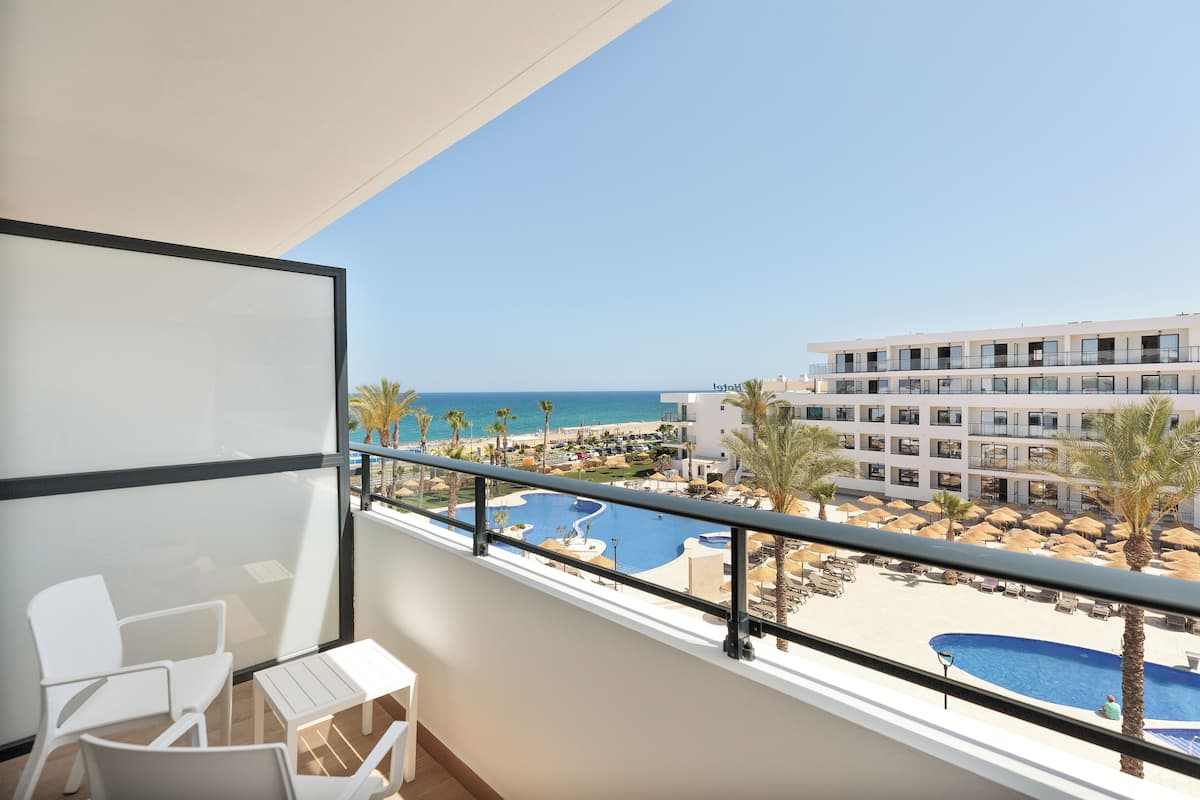 A balcony with white chairs and a small table, overlooking a pool and beach area with palm trees and a hotel building in the background.