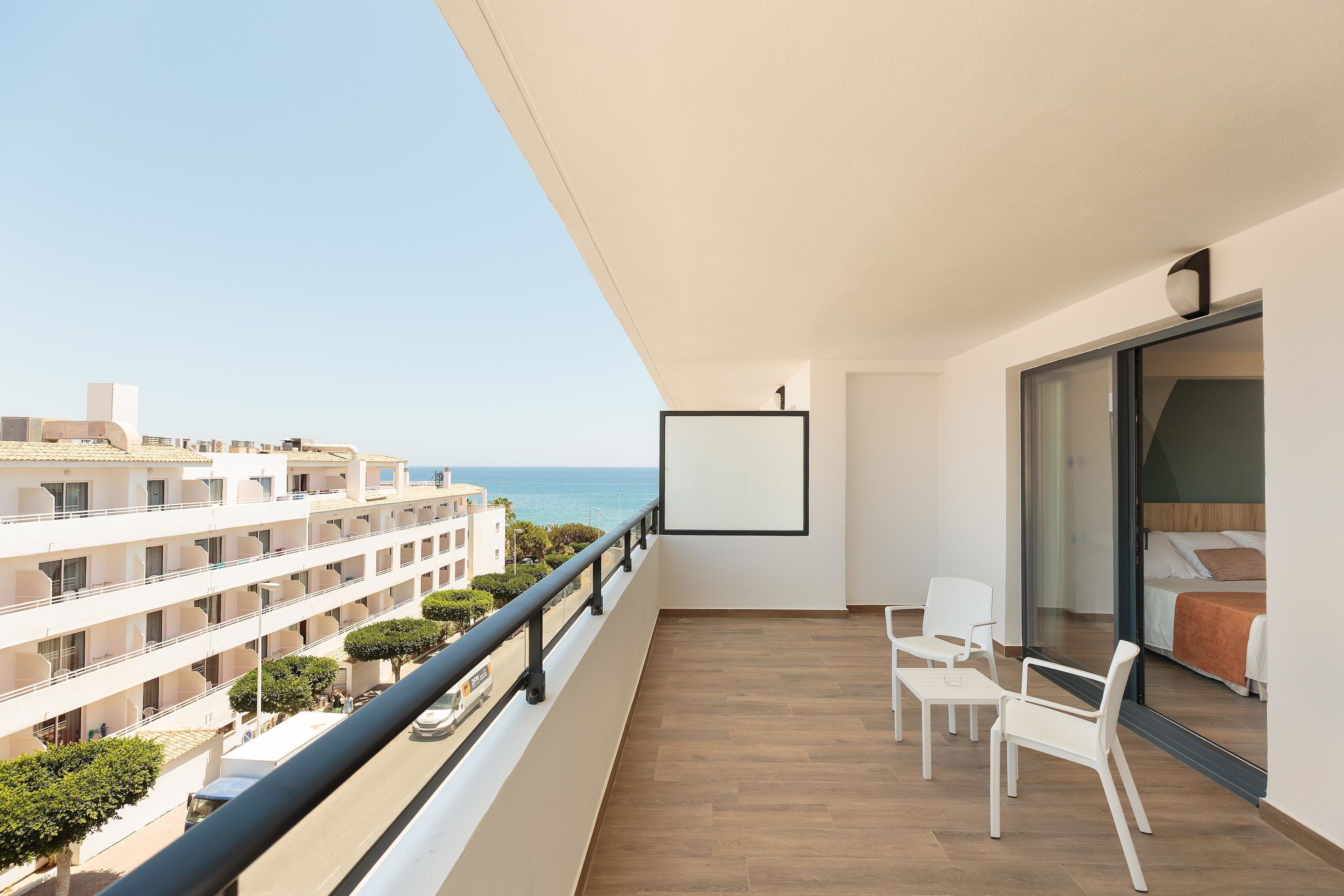 A balcony with a view of the sea, a bed with a brown blanket, and white chairs.