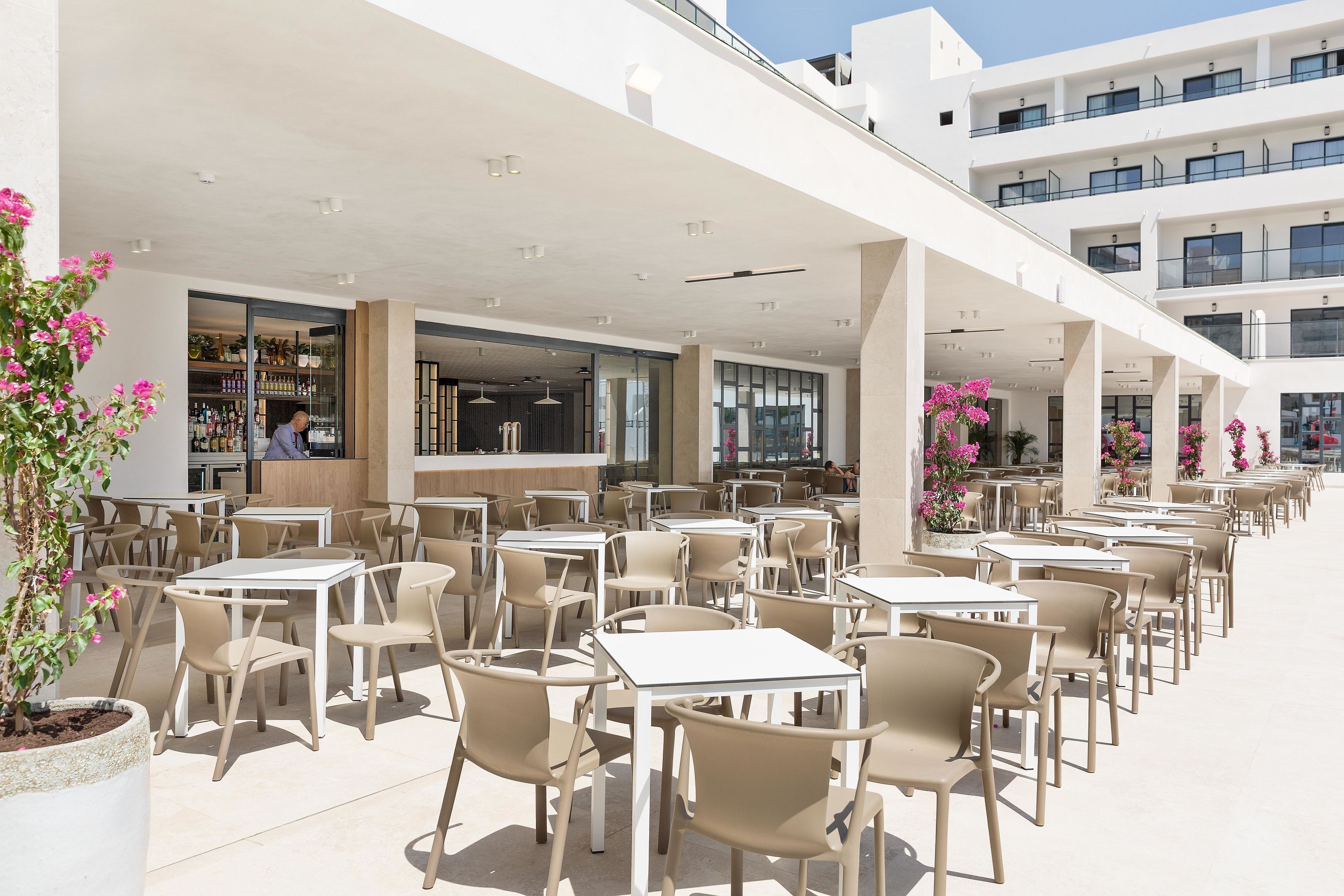Un área de café al aire libre con varias mesas y sillas, una barra visible, y un edificio moderno con balcones al fondo.