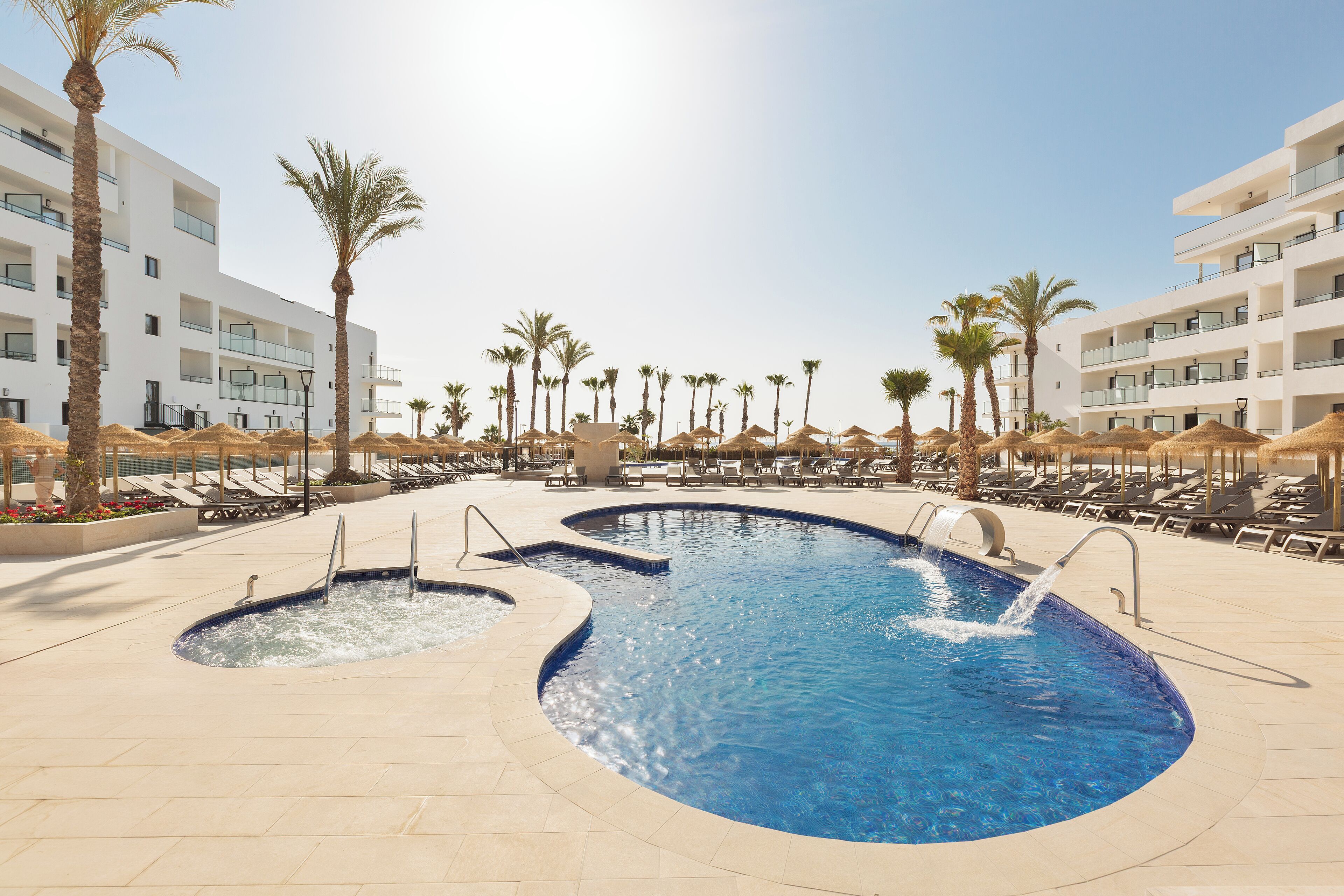 A swimming pool with multiple lanes, surrounded by lounge chairs and palm trees, with a modern building in the background.