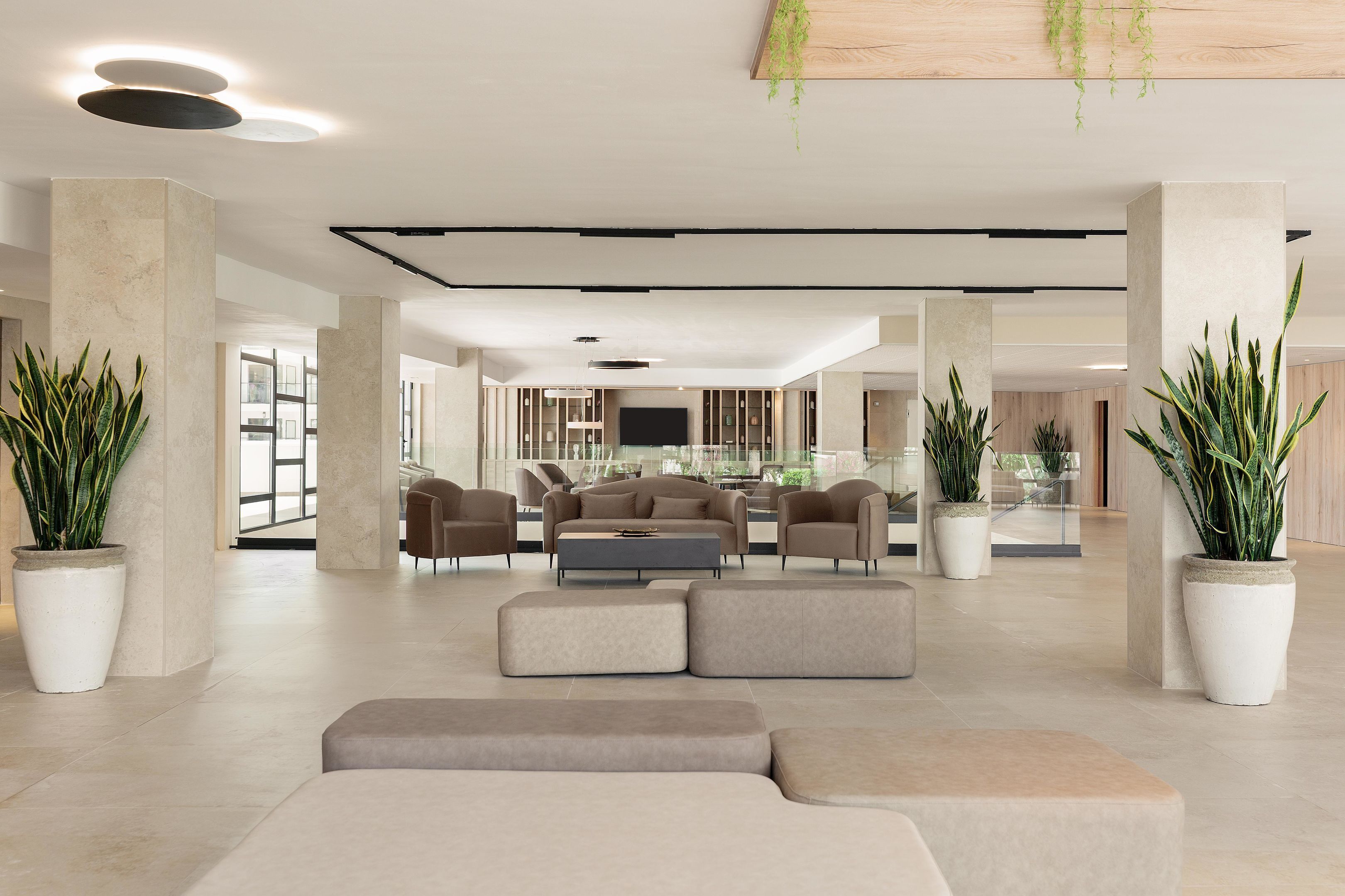 A spacious lobby with a modern design, featuring a central seating area with sofas and armchairs, large potted plants, and a flat-screen TV mounted on the wall.