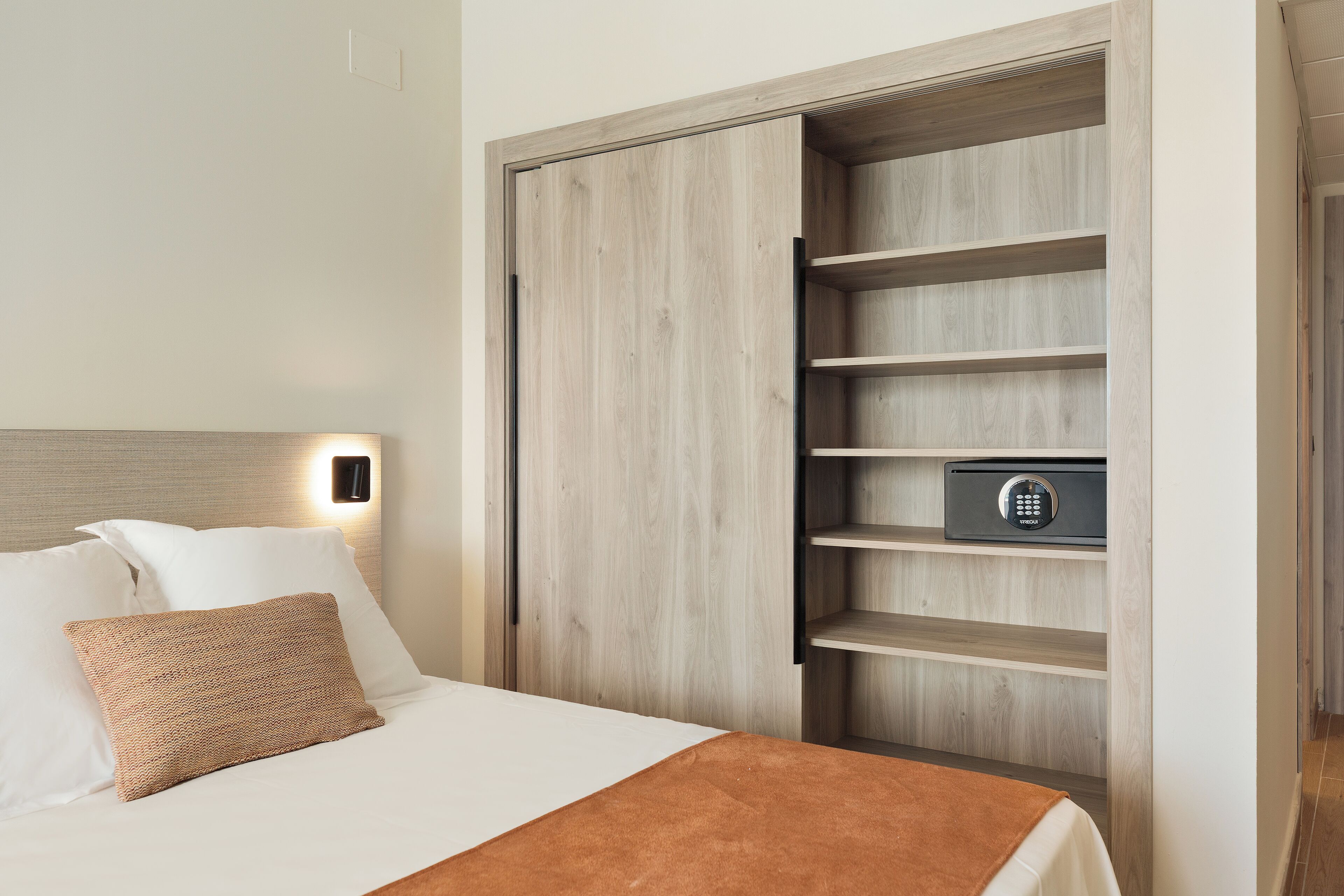 A hotel room with a bed, a wooden wardrobe with shelves, and a safe.