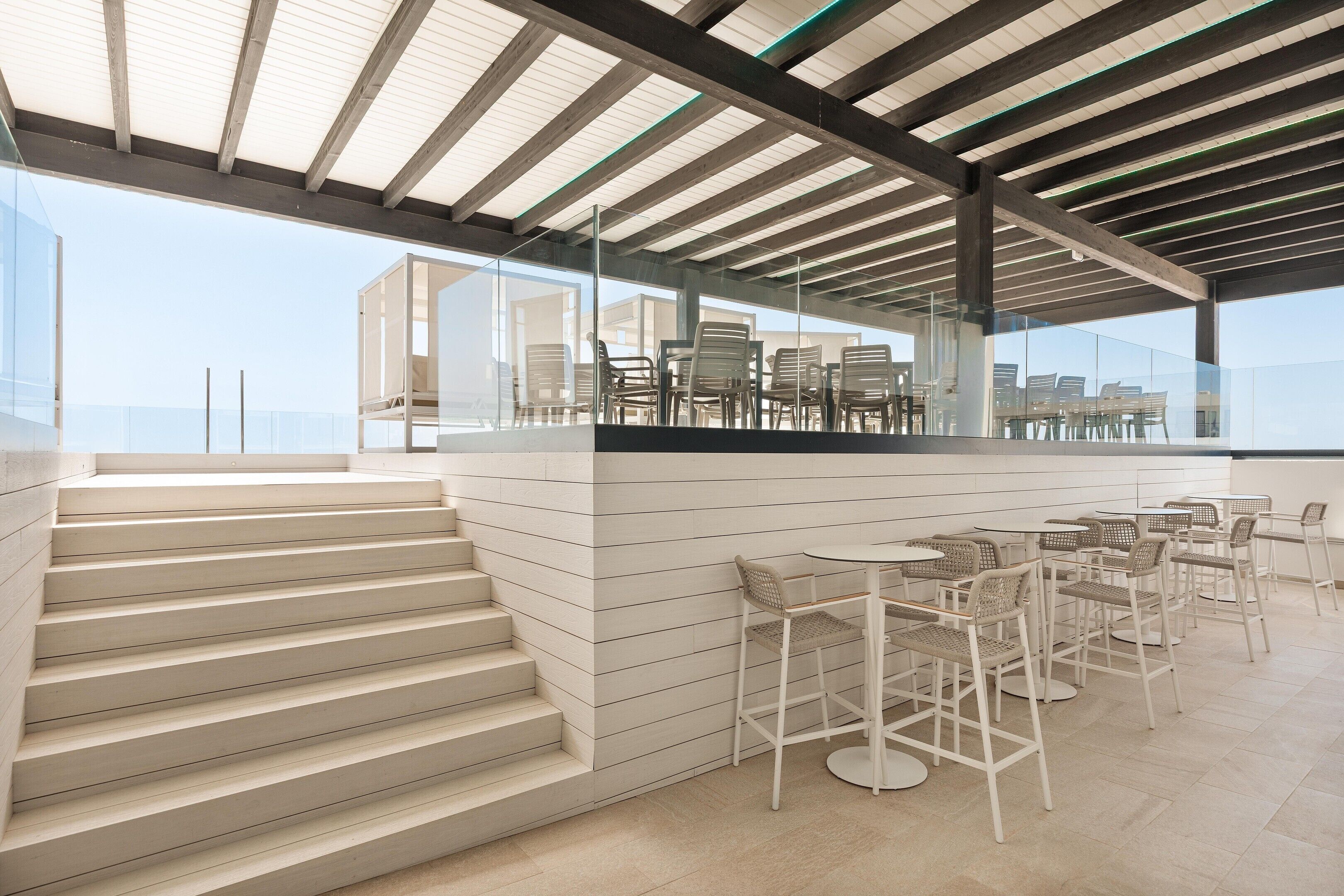 A modern outdoor seating area with a glass railing, white chairs, and a staircase leading to an upper level.