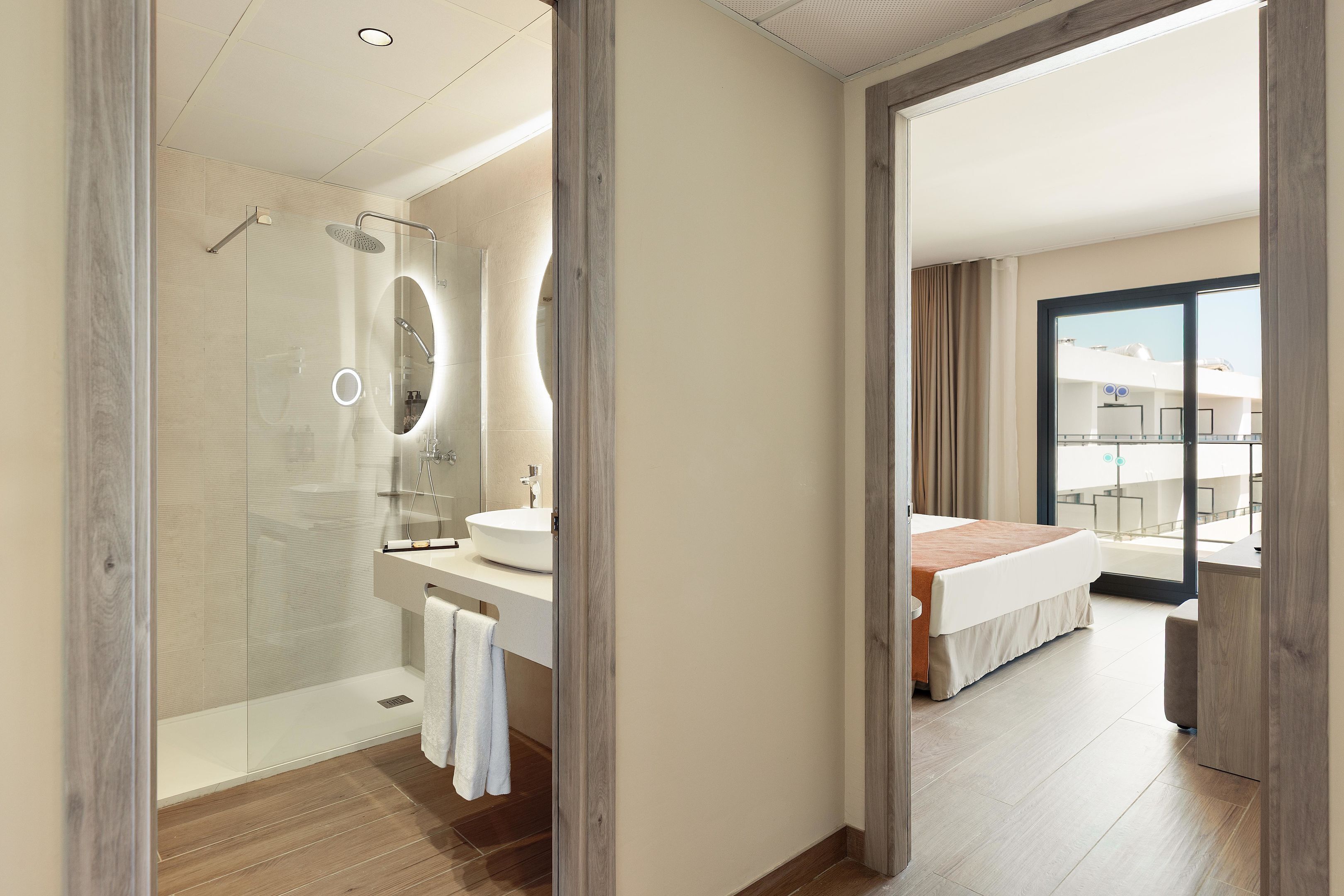 Een moderne hotelkamer met een glazen douche, een grote spiegel, een witte wastafel en een bed met een houten hoofdeinde.