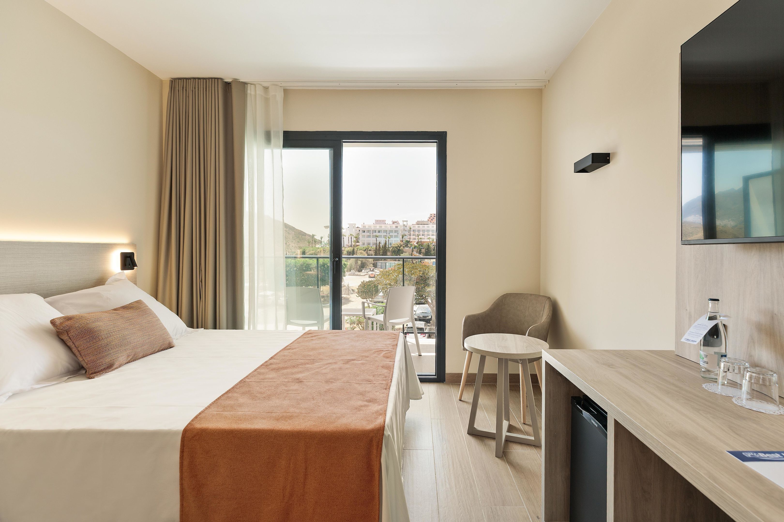 A hotel room with a bed, a small dining area, a chair, a TV, and a view of the city.