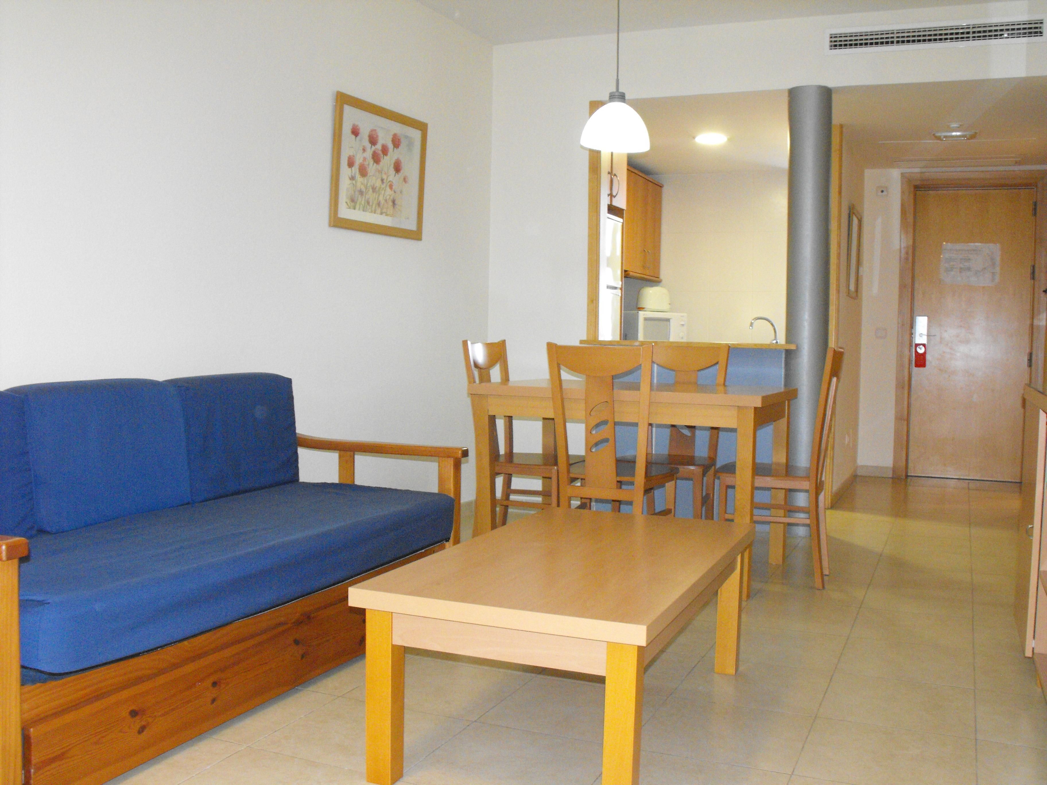 Een kamer met een blauwe bank, een houten eettafel en stoelen. Er is een keukenhoek met een magnetron en kastjes.