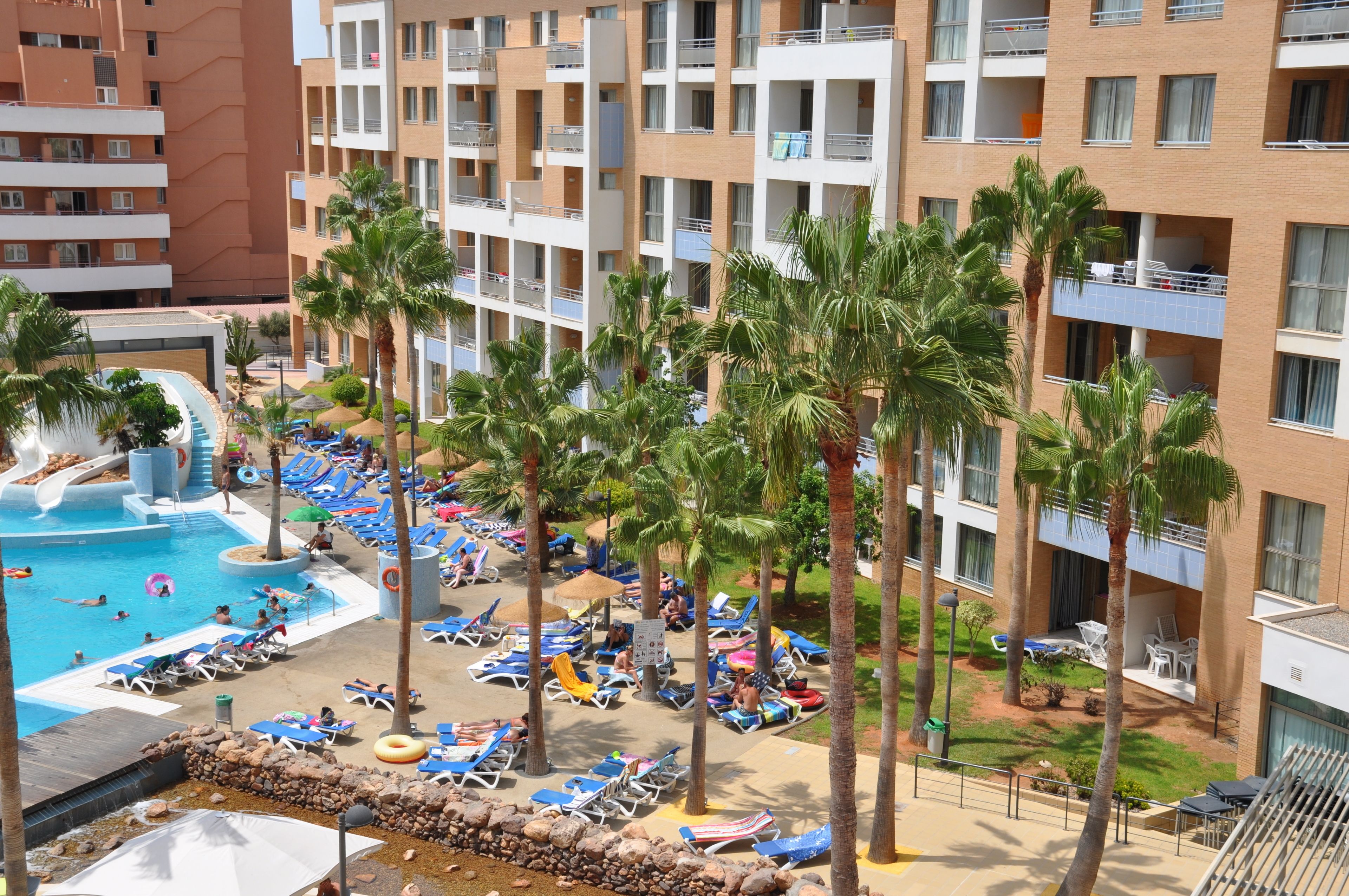 A hotel with a swimming pool, sun loungers, palm trees, and a water slide.