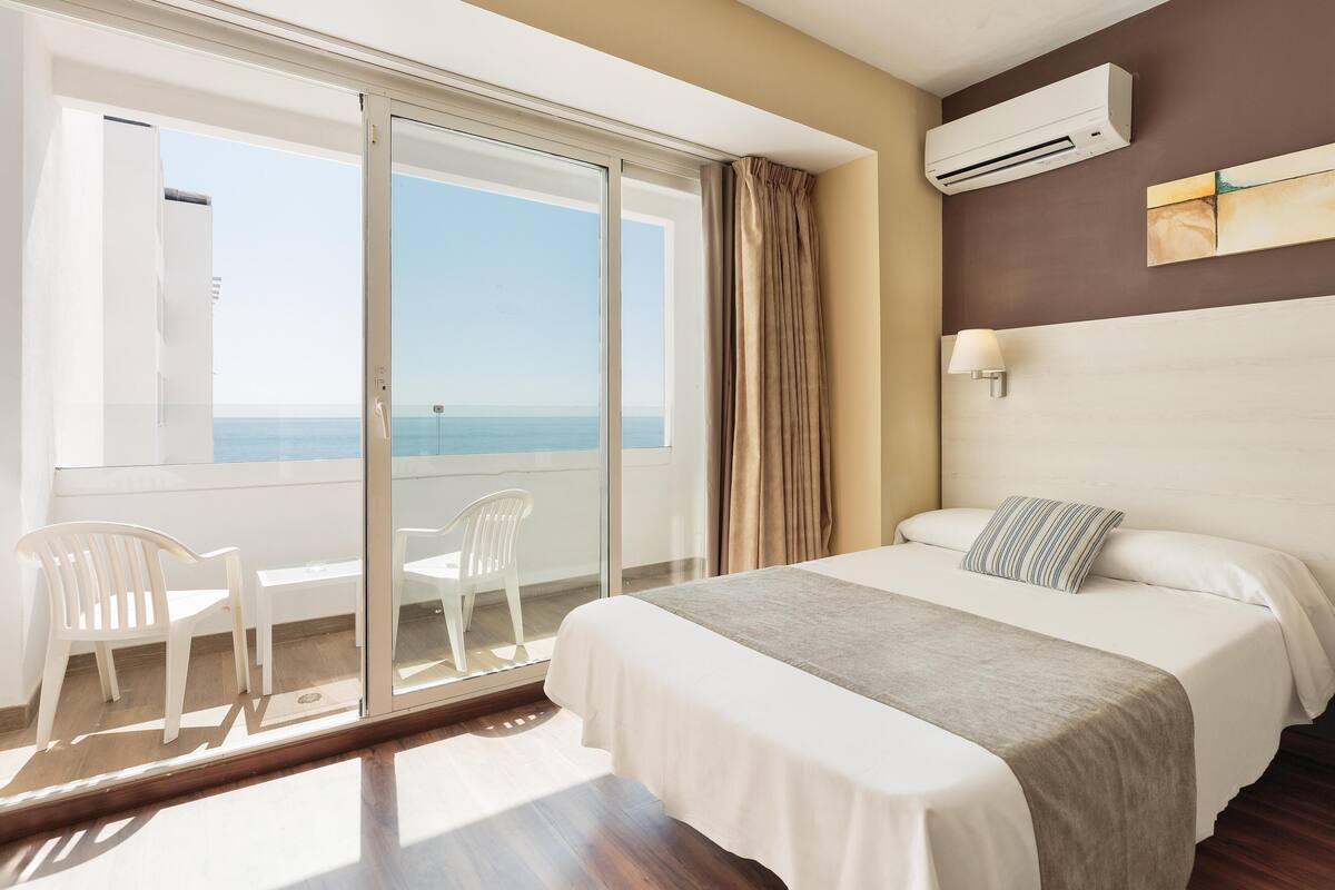 A hotel room with a bed, a view of the sea, a balcony with chairs, and an air conditioning unit.