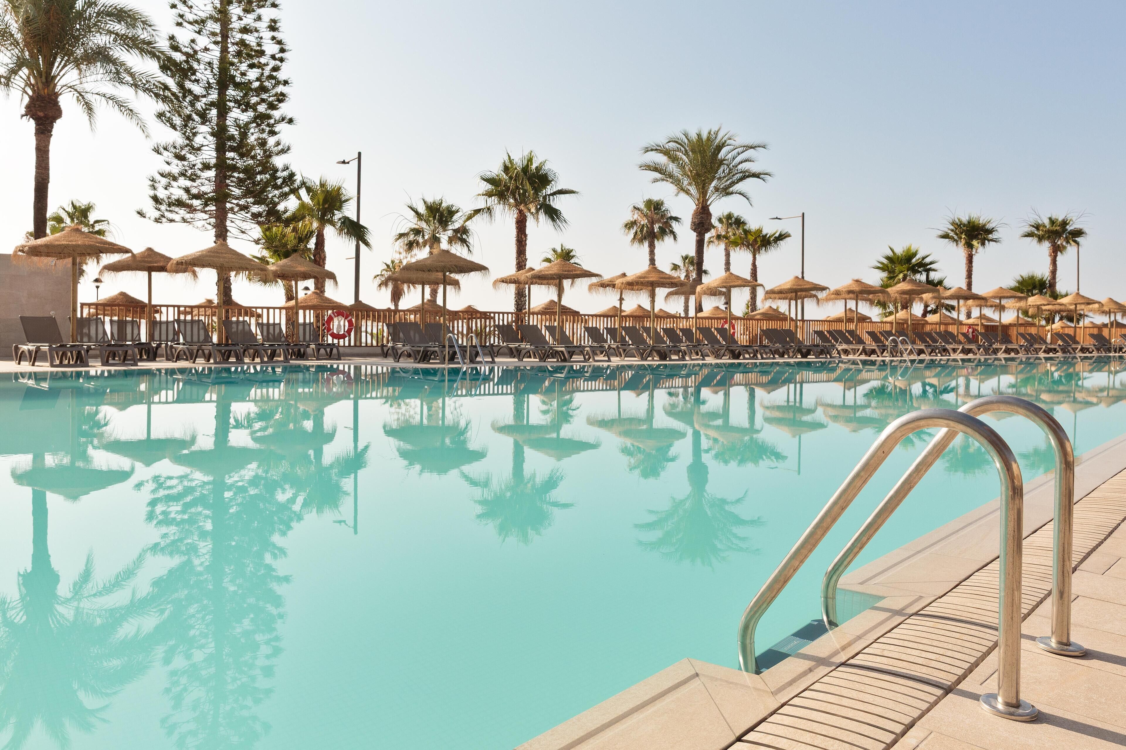 Zona de piscina con sillones de descanso y sombrillas, rodeada de palmeras.