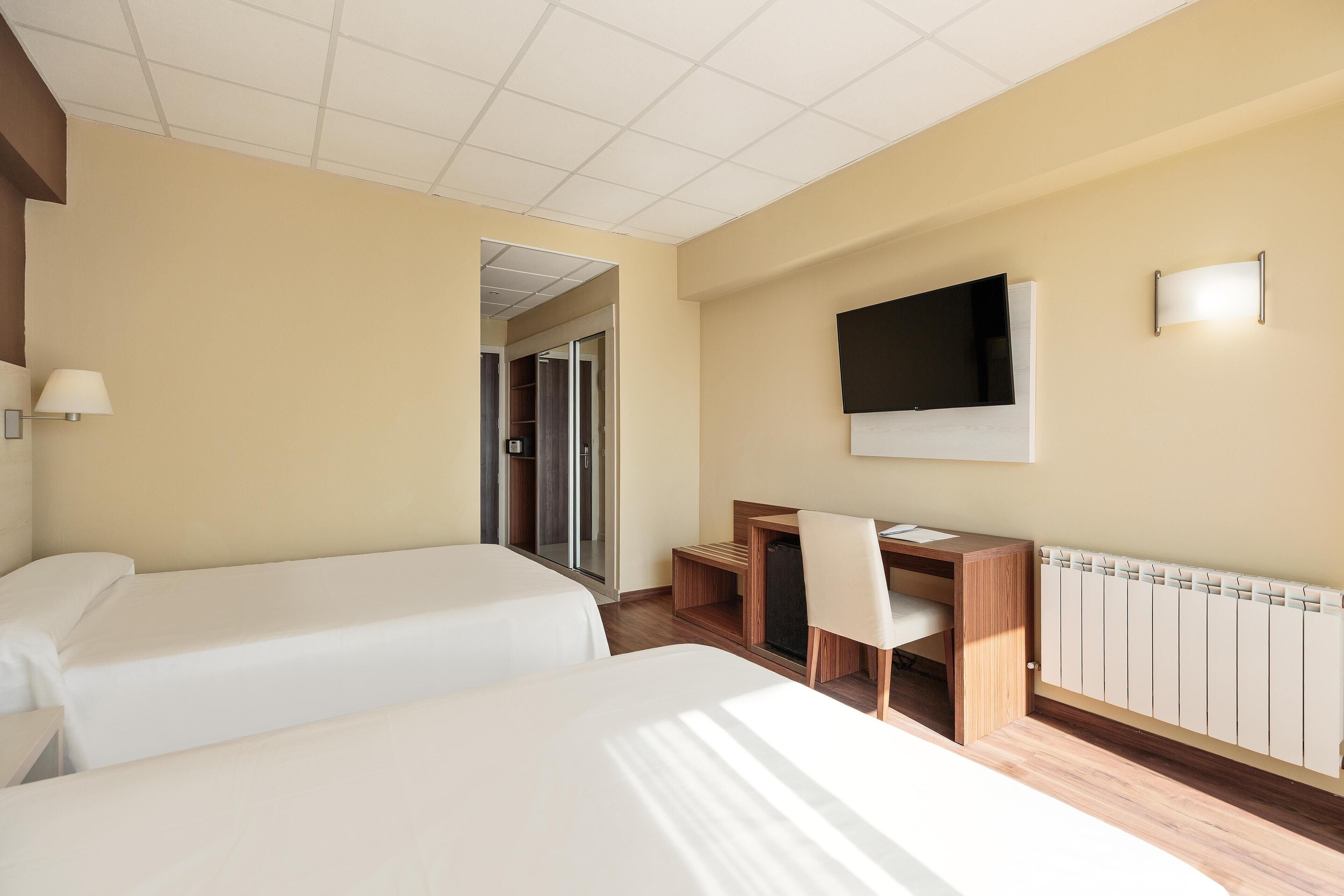 A hotel room with two beds, a desk, a chair, a TV, and a window.