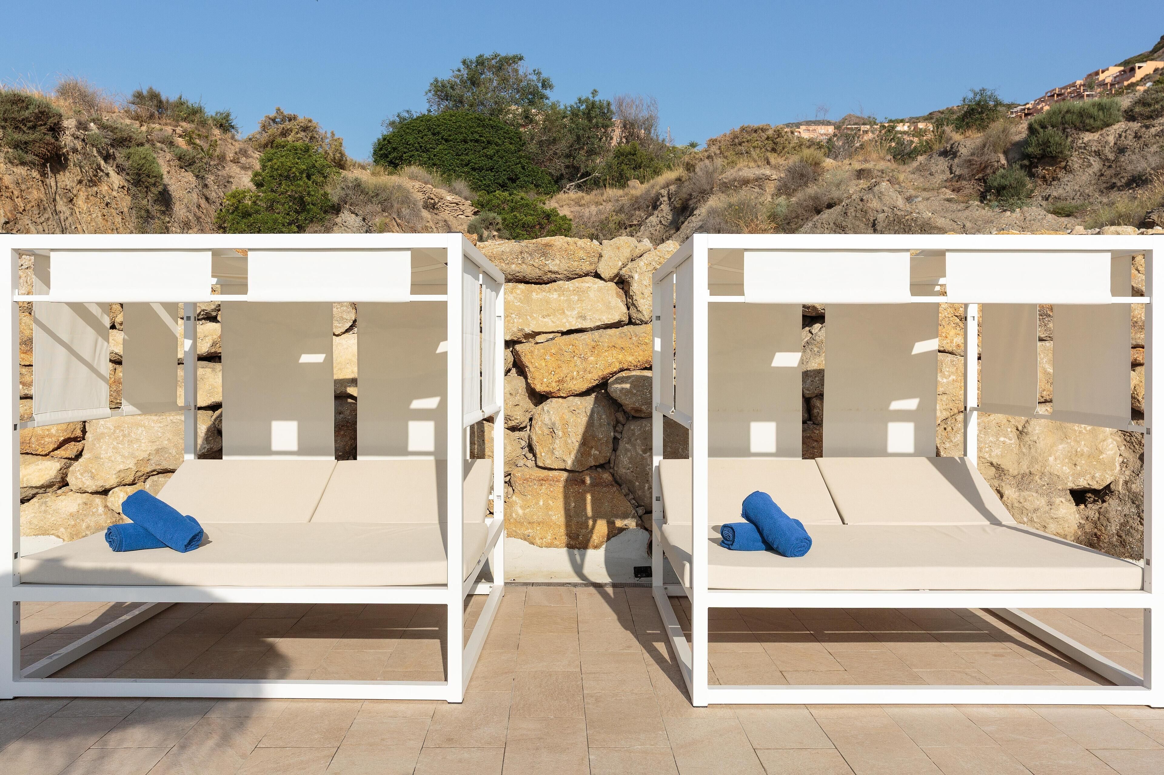 Two white outdoor lounge chairs with cushions, set against a backdrop of stone walls and a clear sky.