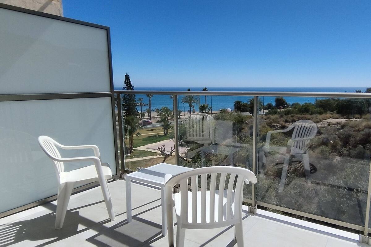 A balcony with two white plastic chairs and a small table, overlooking a coastal area with a clear view of the sea and greenery.