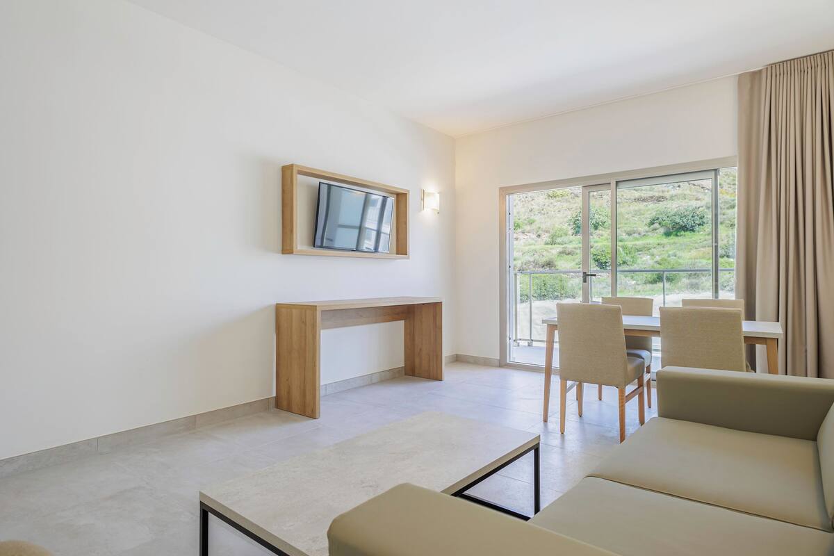 A modern living room with a sofa, a coffee table, a TV mounted on the wall, and a dining area with chairs and a table.