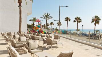 A rooftop terrace with wicker furniture, palm trees, and a view of the beach.