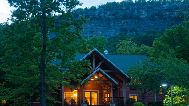 A lodge with a prominent front porch, a stone staircase leading up to the entrance, and a view of a rocky cliff in the background. - Minnewaska Lodge (Gardiner)