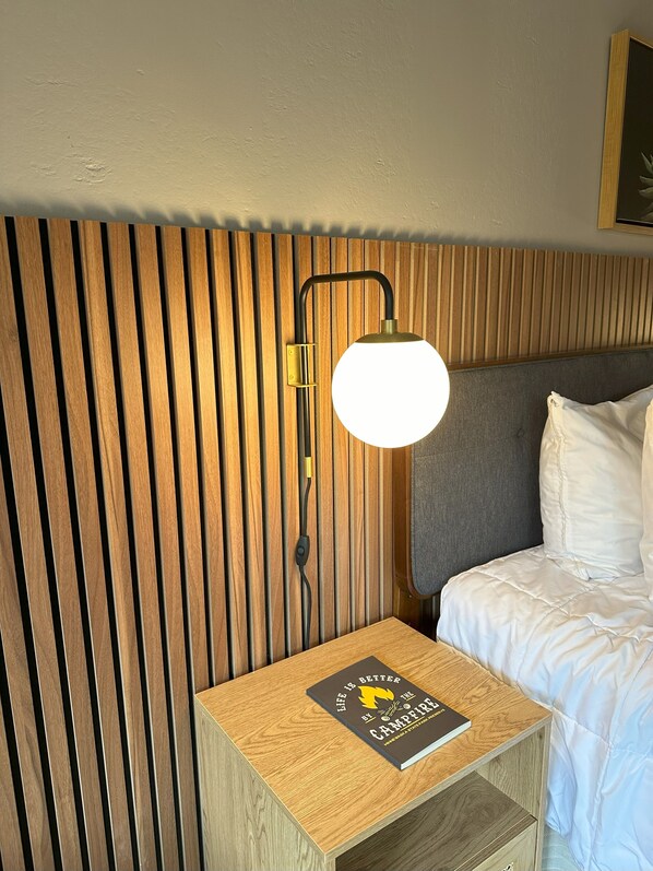 A neatly made bed with white linens, a wooden bedside table with a book titled "Life is Better by the Campfire," and a modern wall-mounted lamp. - Minnewaska Lodge (Gardiner)