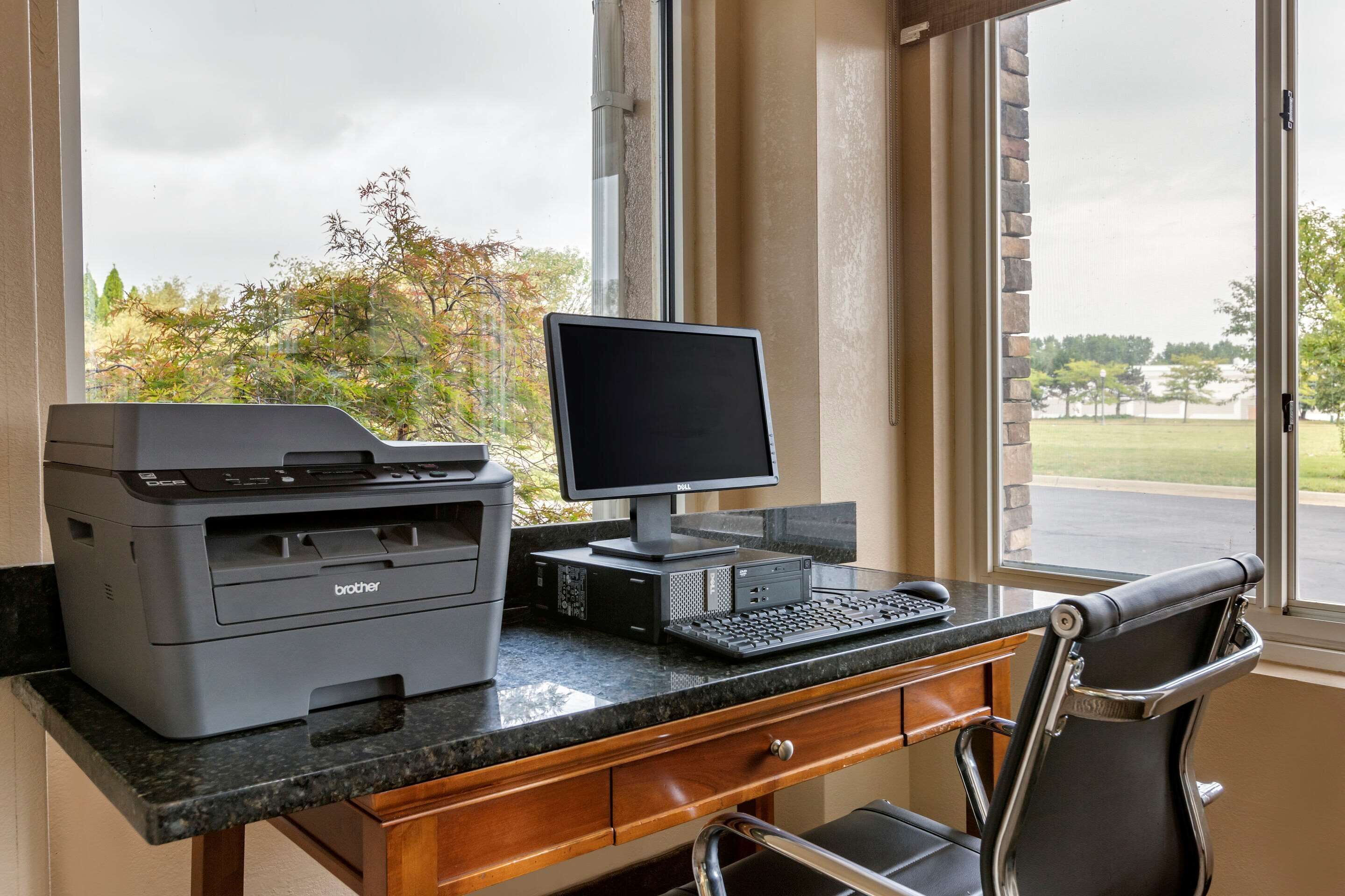 Um espaço de escritório em casa com computador de mesa, impressora e cadeira próximo a uma janela com vista para árvores e um estacionamento.