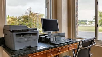 Um espaço de escritório em casa com computador de mesa, impressora e cadeira próximo a uma janela com vista para árvores e um estacionamento.