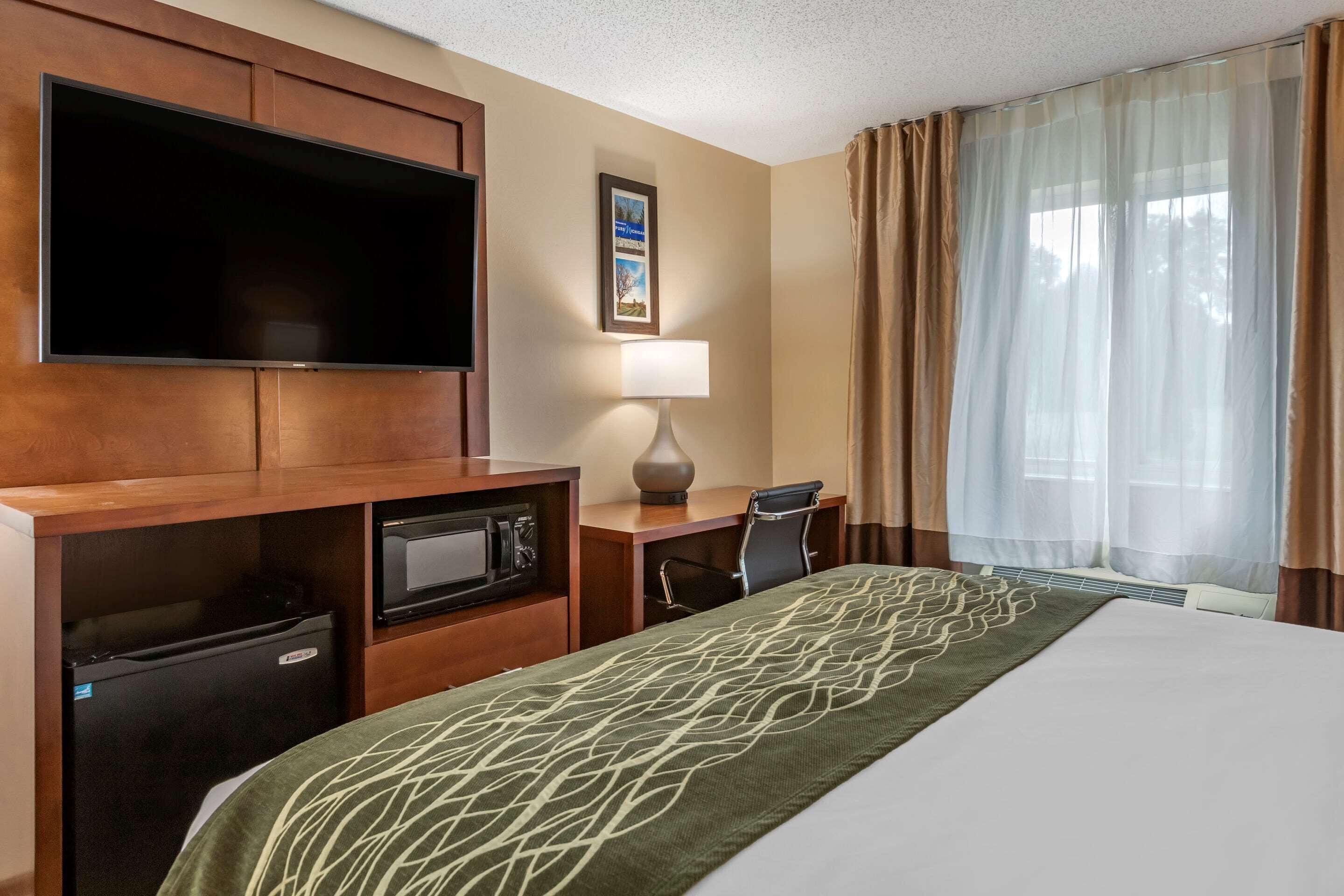Habitación de hotel con cama, escritorio con computadora, televisión, lámpara y ventana con cortinas.