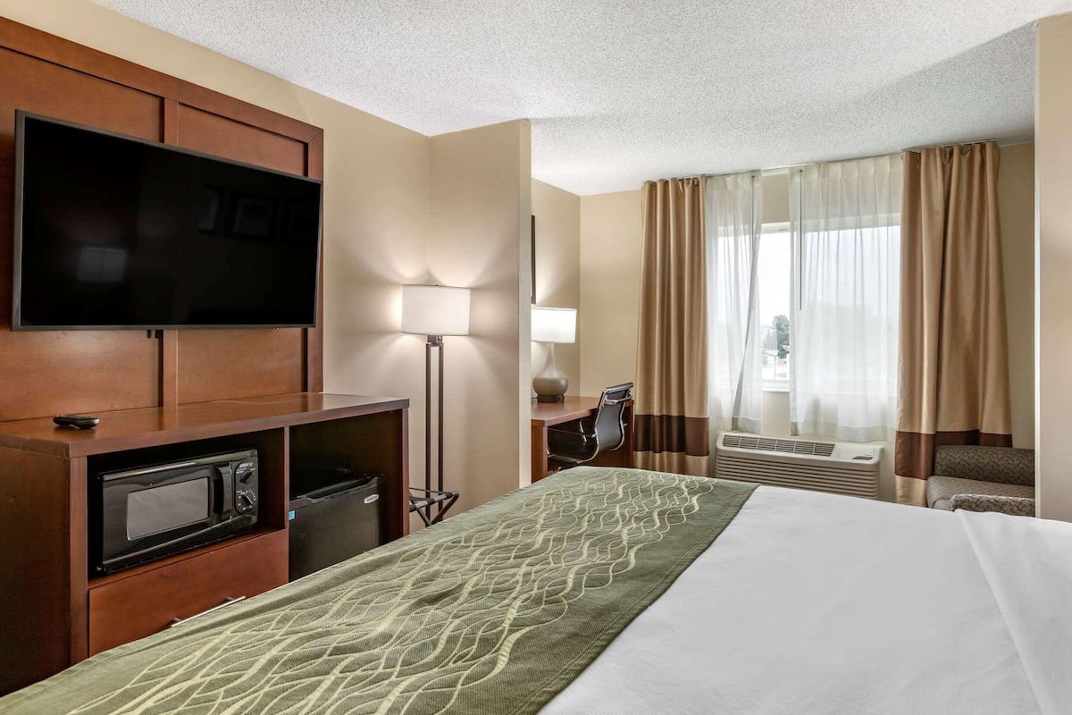 Une chambre d’hôtel comprenant un lit, un téléviseur à écran plat fixé à une armoire en bois, un four à micro-ondes, un bureau avec une lampe et une grande fenêtre avec des rideaux.