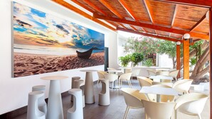 Un espace extérieur moderne aménagé avec des chaises et des tables blanches originales, une grande peinture représentant un bateau sur une plage et une structure de plafond en bois.
