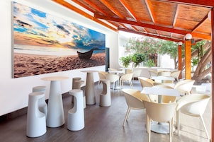Un espacio moderno al aire libre con sillas y mesas blancas de diseño, un cuadro grande de un bote en la playa, y una estructura de techo de madera.