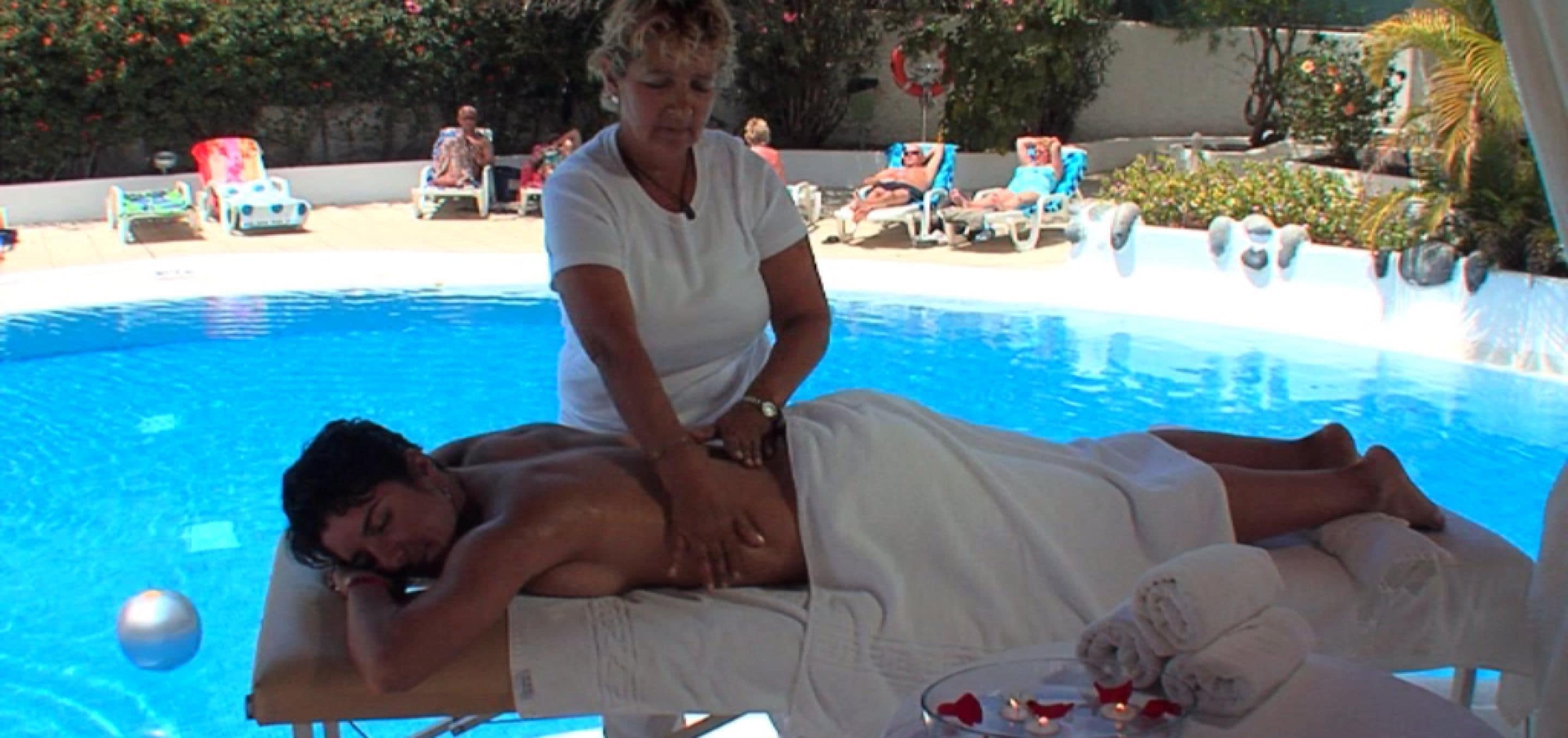 Un hombre recibe un masaje de una mujer en un entorno junto a la piscina, con sillones de descanso y una piscina al fondo.