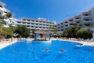 Una piscina con un pabellón, rodeada de sillas de descanso y palmeras, con un edificio de hotel al fondo.