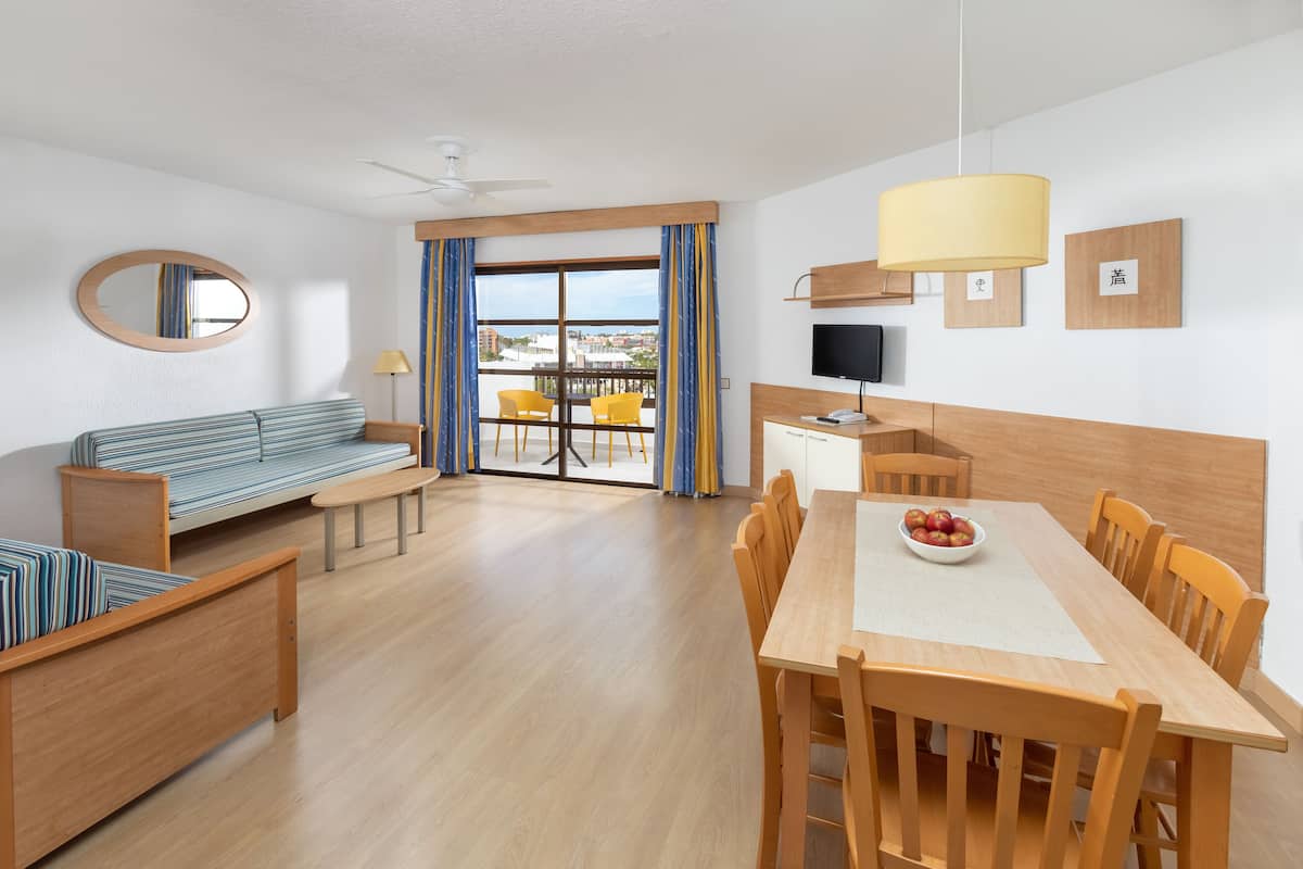 Habitación de hotel moderna con una mesa de comedor de madera, un sofá, un televisor y un balcón con sillas amarillas.
