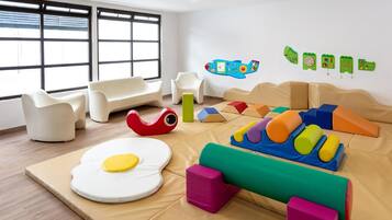 A modern playroom with a large window, white sofas, and colorful play structures.