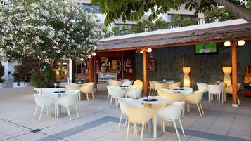 Un café en plein air avec des chaises blanches et jaunes, un toit de chaume et un écran de télévision fixé au mur.