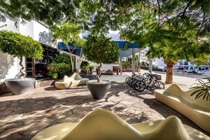 Un espacio al aire libre con sillas modernas y macetas, una bicicleta estacionada cerca, y un edificio con techo azul al fondo.