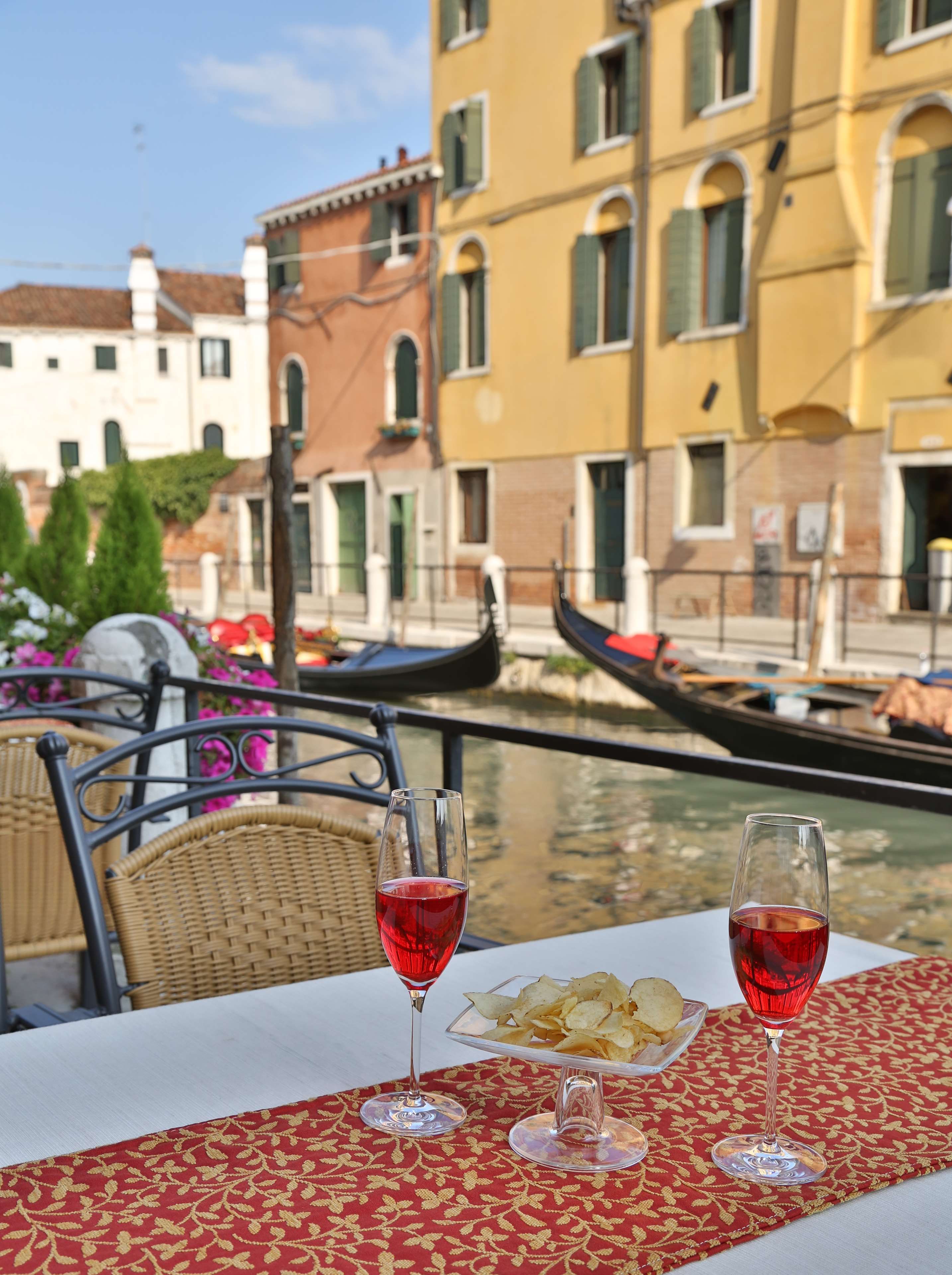 A table with two wine glasses and a plate of chips, set against a backdrop of a canal with gondolas and buildings.