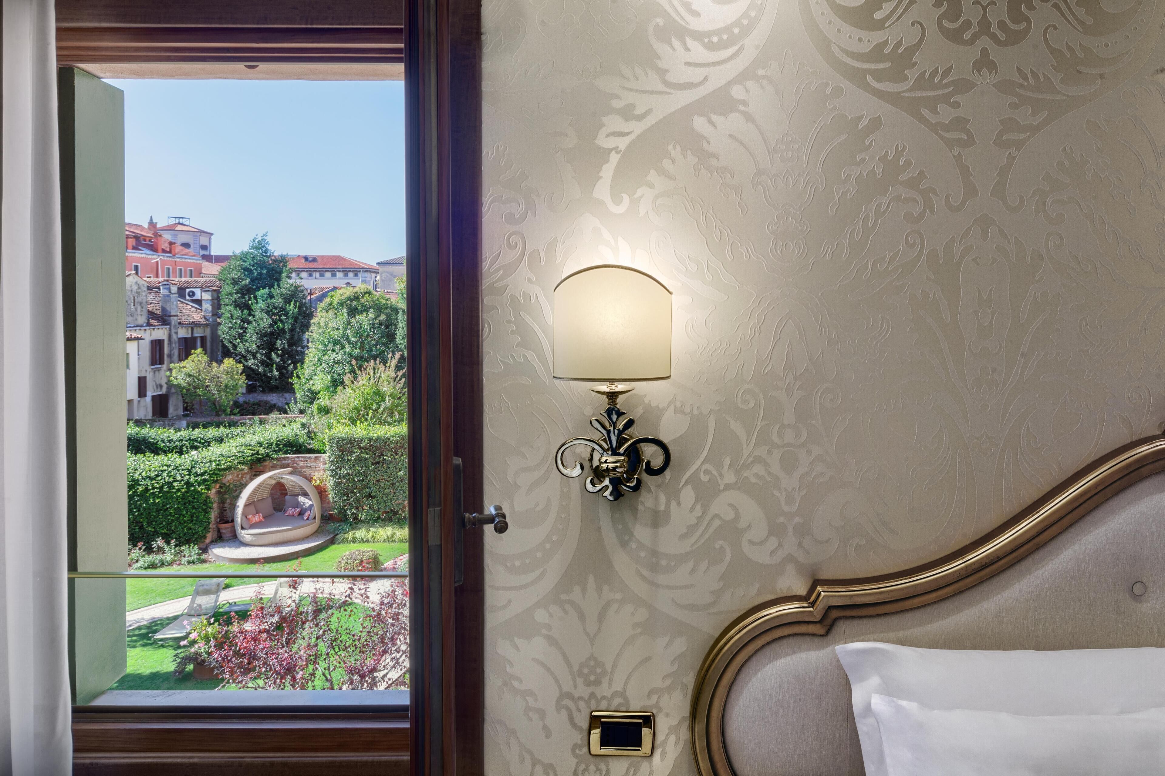 Habitación de hotel con vistas a un jardín y edificios. Incluye una cama con un almohadón blanco, una lámpara fijada a la pared y un pequeño dispositivo cuadrado en la pared.