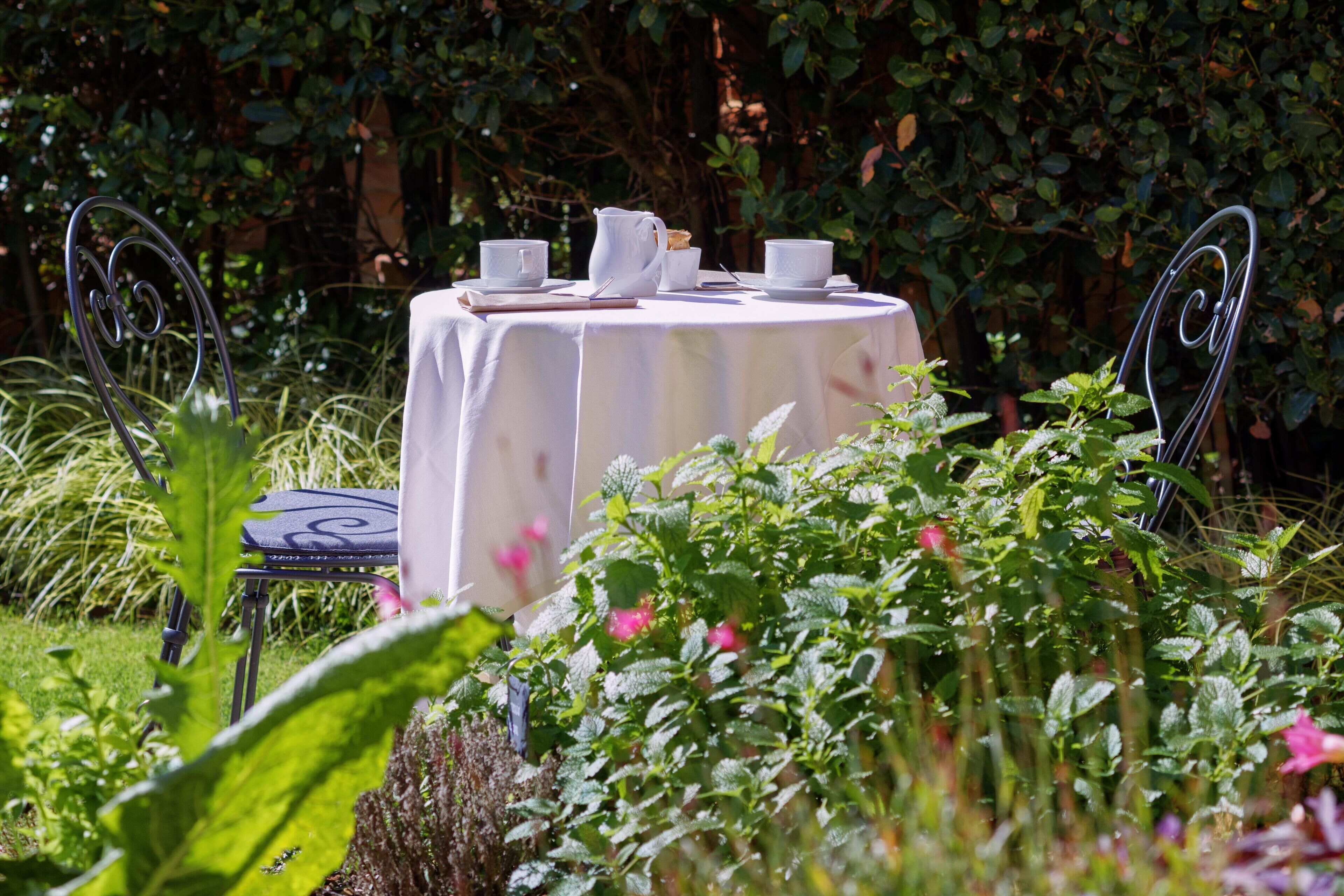 A round table with a white tablecloth, surrounded by metal chairs, set in a garden with lush greenery and flowering plants.