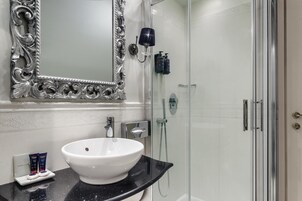 Um banheiro com pia branca, um espelho grande com moldura prateada ornamentada, bancada preta e área de chuveiro com divisória de vidro.