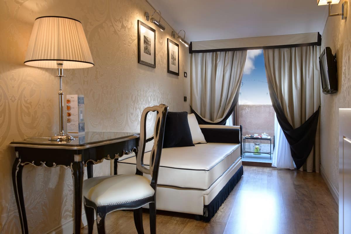 A compact hotel room with a bed, a desk with a lamp, a chair, and a small table with a cup and saucer.