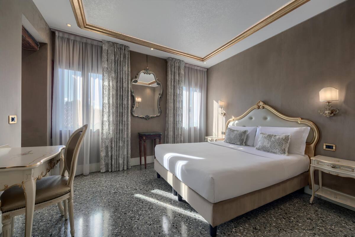 A hotel room with a large bed, a desk, and a chair. There is a mirror on the wall and a lamp on the bedside table.