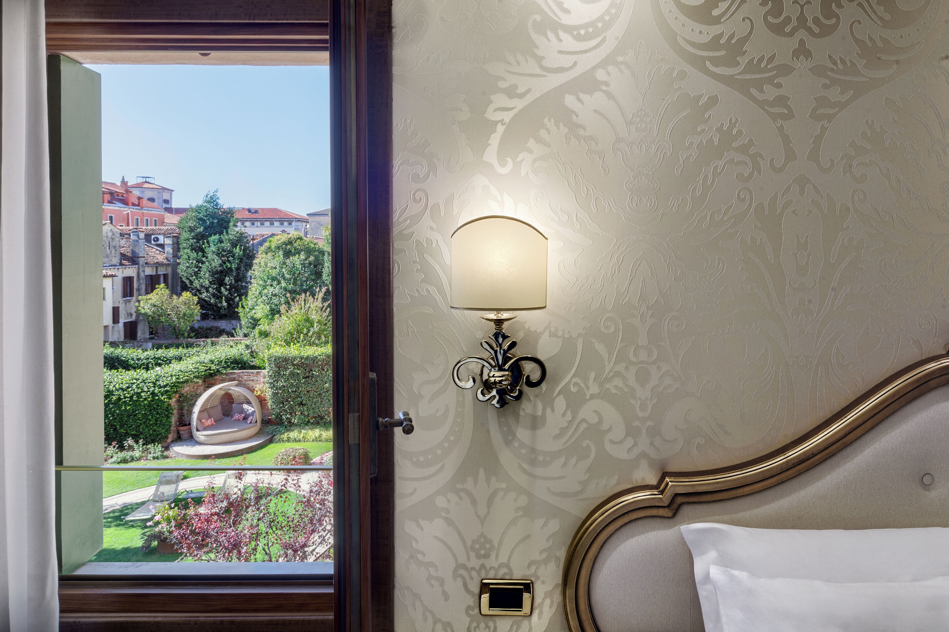 A hotel room with a view of a garden and buildings. It includes a bed with a white pillow, a wall-mounted lamp, and a small square object on the wall.
