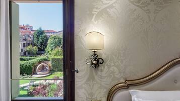 A hotel room with a view of a garden and buildings. It includes a bed with a white pillow, a wall-mounted lamp, and a small square object on the wall.