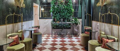 A lobby with a checkered floor, green upholstered chairs, a central planter with plants, and decorative wall sconces.