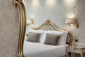 A bedroom with a large ornate mirror, a bed with white linens and two pillows, two wall-mounted lamps, and a nightstand with a glass top. - Hotel Olimpia Venice, BW Signature Collection (Venice)