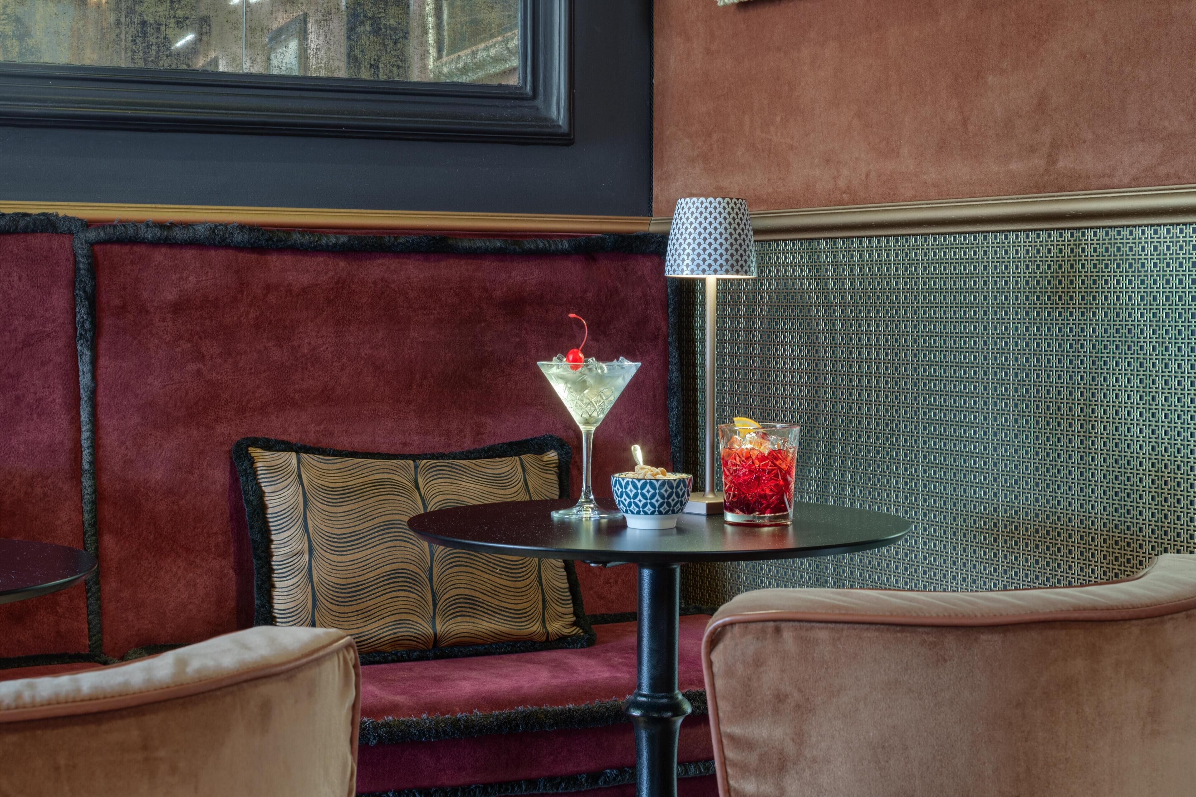 A cozy seating area with a red velvet sofa, a patterned cushion, a small round table with a martini glass and a bowl, and a patterned lamp.
