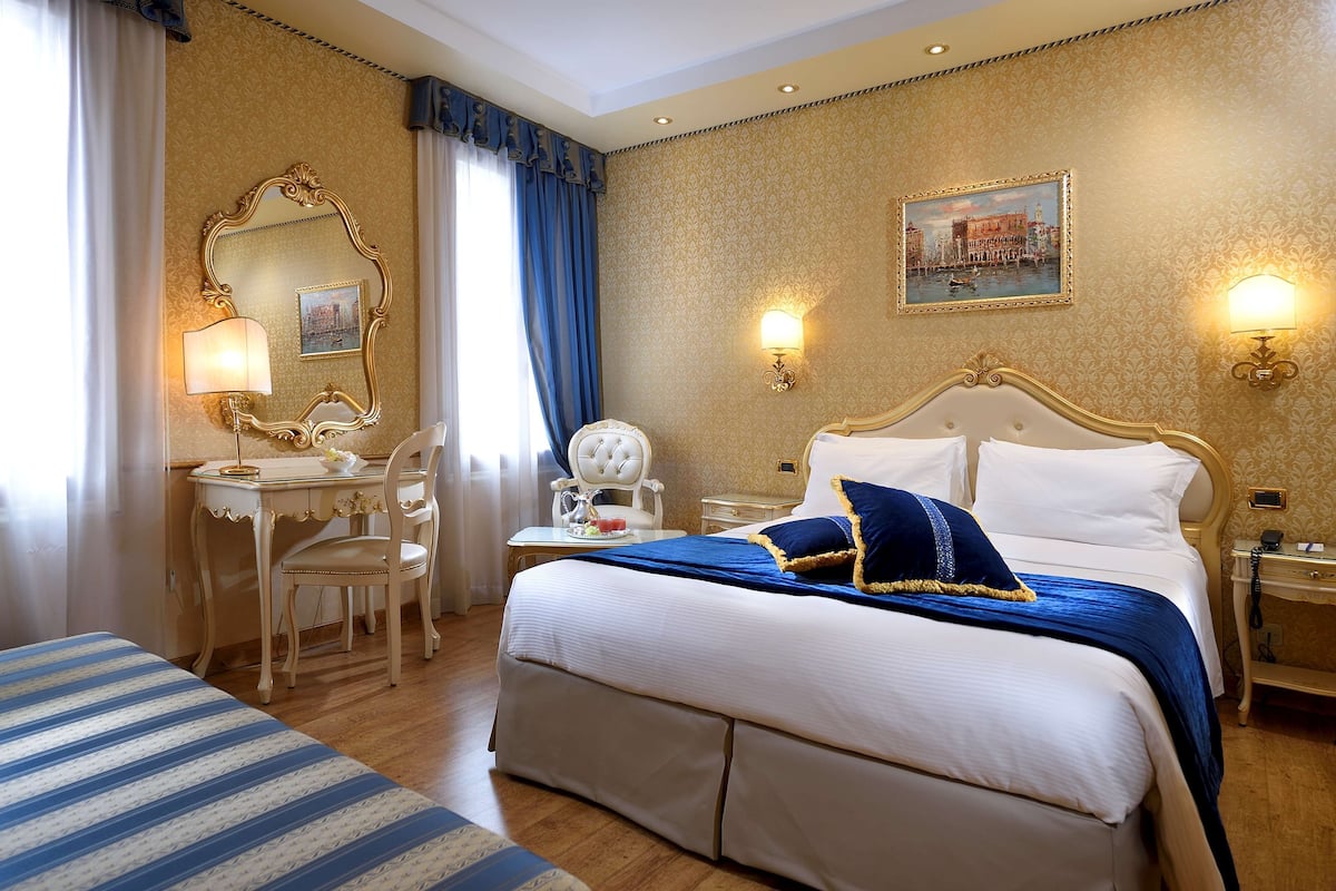 A hotel room with a large bed, a desk with a chair, a small table with a lamp, and a painting on the wall.