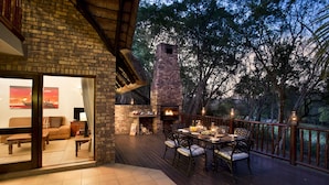 A deck with a brick fireplace, outdoor seating, and a view of trees. - Kruger Park Lodge (Mbombela)
